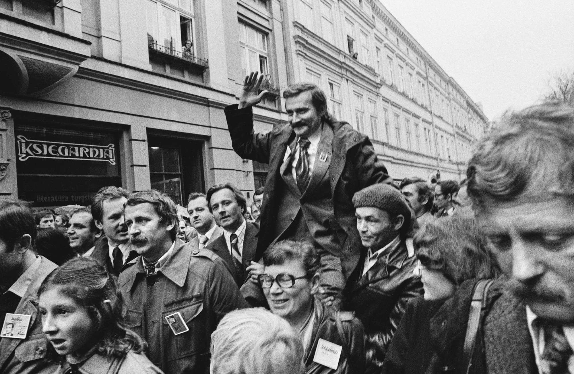 Lech Wałęsa mit Anzug und Krawatte wird von einer Menschenmenge, die durch eine Straße zieht, auf Schultern getragen. Er winkt mit der rechten Hand. (Schwarz-weiß-Aufnahme)