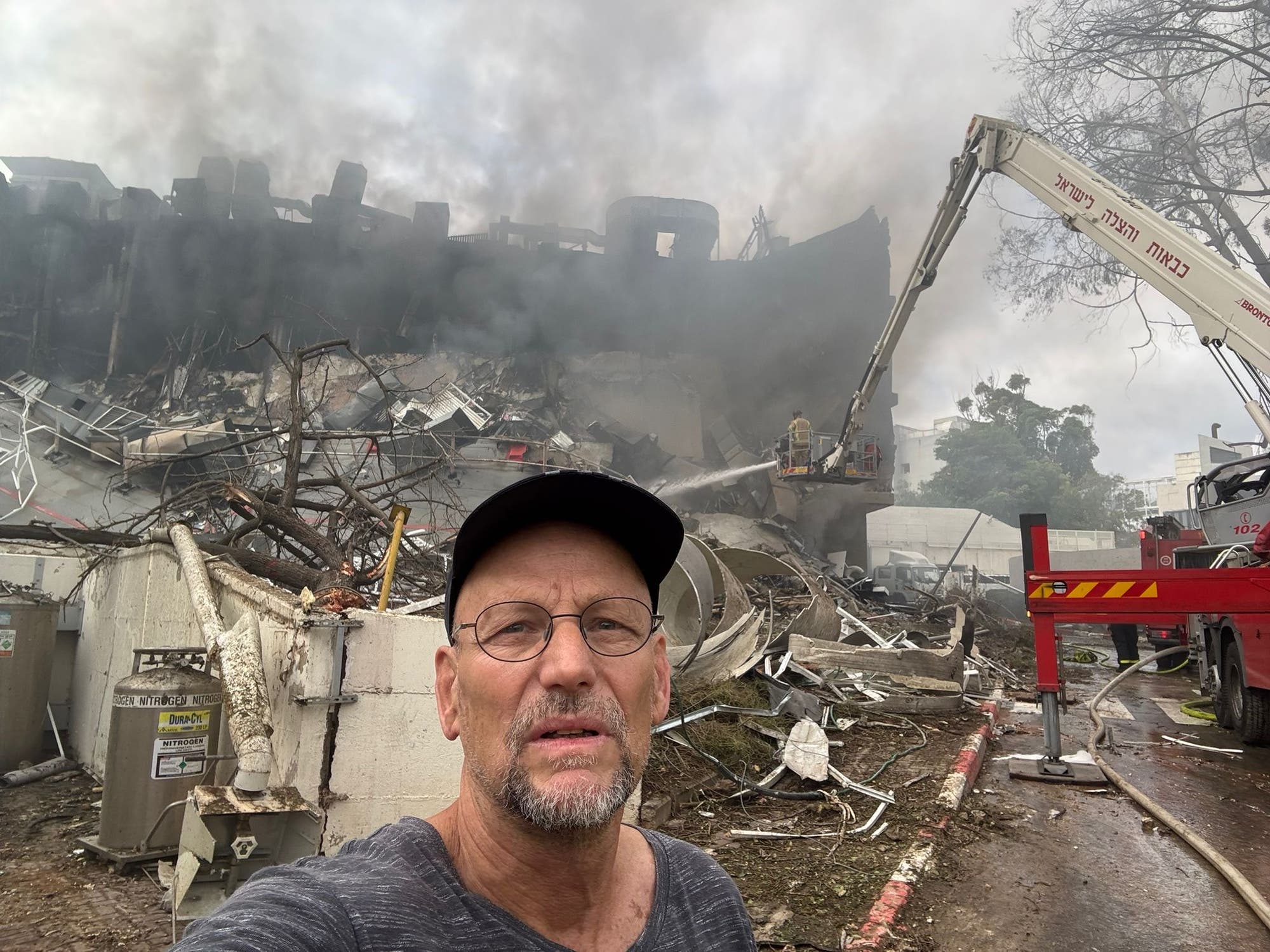 Eine Person mit Brille und Mütze steht vor einem zerstörten Gebäude, aus dem Rauch aufsteigt. Im Hintergrund ist ein Kran zu sehen, der an den Aufräumarbeiten beteiligt ist. Trümmer und Schutt sind überall verstreut. Die Szene vermittelt den Eindruck von Zerstörung und Wiederaufbauarbeiten.