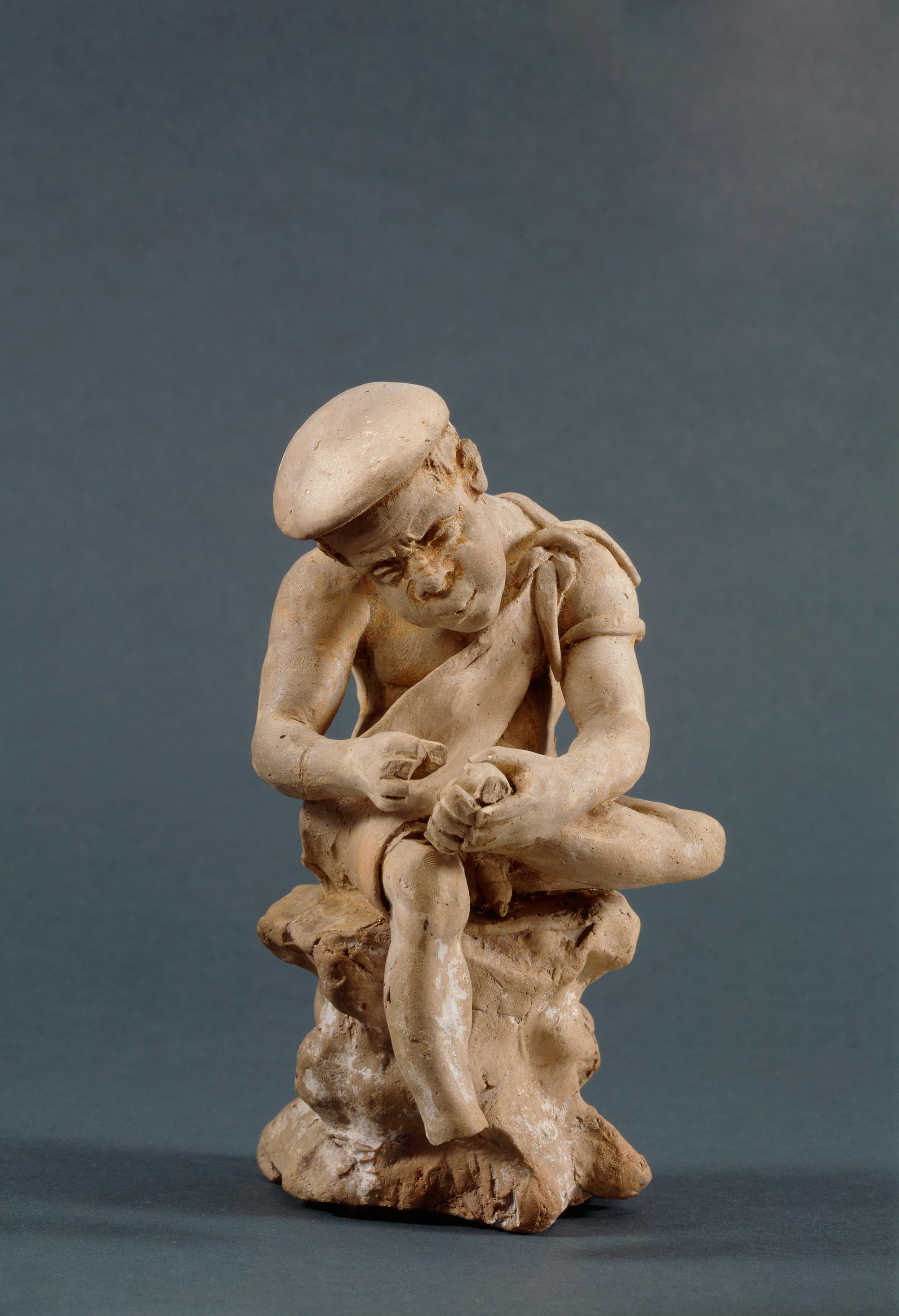Eine Terrakotta-Skulptur eines sitzenden Mannes, der einen Dorn aus seinem Fuß zieht. Der Mann trägt eine Mütze und ein einfaches Gewand, während er auf einem Felsen sitzt. Der Hintergrund ist neutral grau, was den Fokus auf die detaillierte Darstellung der Figur lenkt.