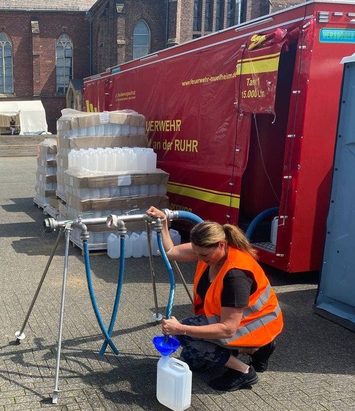 Eine Frau in einer orangefarbenen Warnweste füllt Wasser aus einem mobilen Wasserhahn in einen Kanister. Im Hintergrund steht ein rotes Feuerwehrfahrzeug mit der Aufschrift "Feuerwehr Mülheim an der Ruhr" und "Tank 15.000 Ltr.". Daneben sind gestapelte leere Kanister zu sehen. Die Szene spielt sich vor einem großen Gebäude mit gotischen Fenstern ab. Die URL auf dem Fahrzeug lautet "www.feuerwehr-muelheim.de".