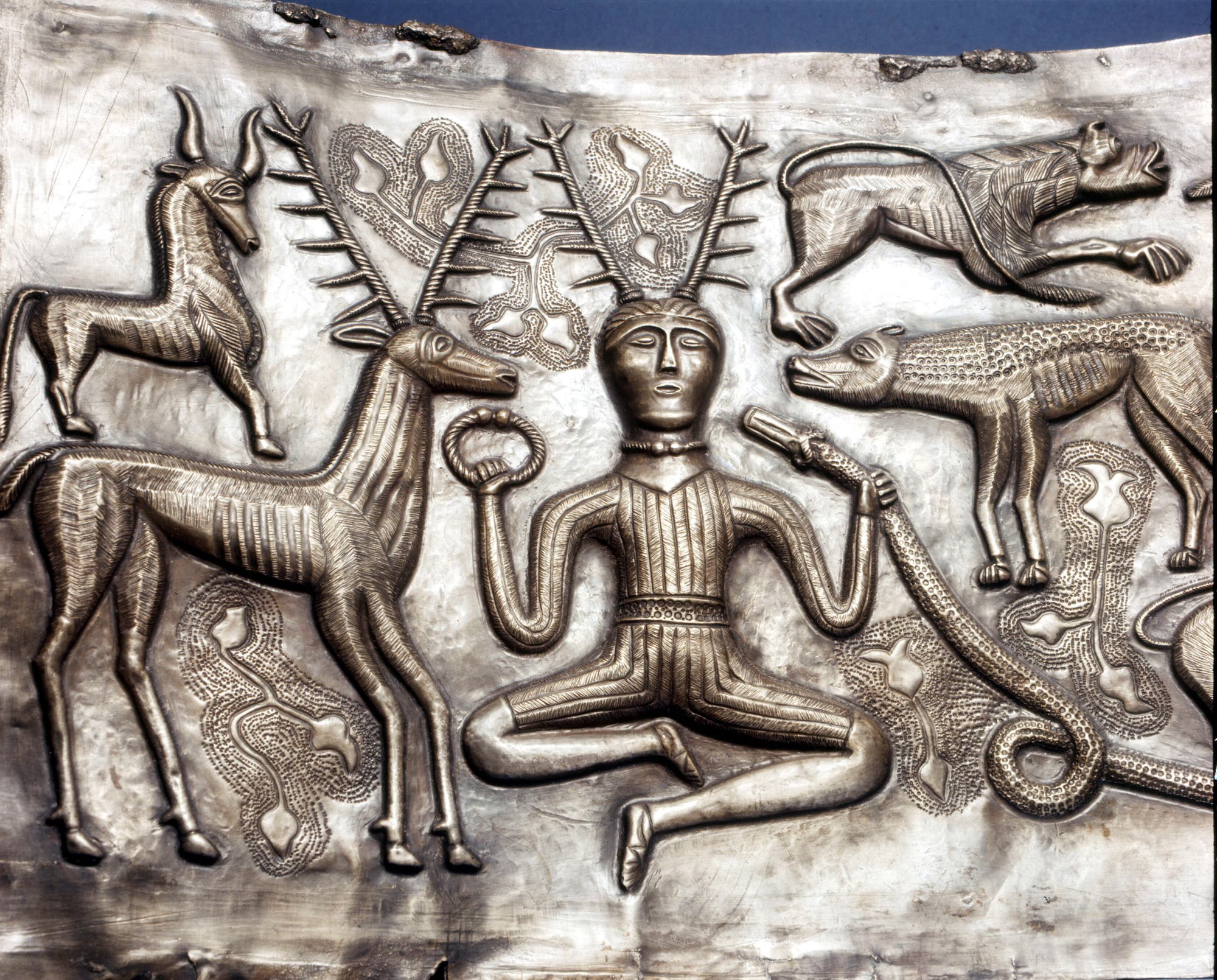 Der Ausschnitt einer antiken, kunstvoll gestalteten metallenen Schale zeigt eine zentrale Figur mit Geweih, die im Schneidersitz sitzt. Die Figur hält in der rechten Hand einen Ring und in der linken eine Schlange. Um die Figur herum sind verschiedene Tiere abgebildet, darunter eine Hirschkuh und womöglich eine Ziege, ein Löwe und ein Wolf. Die Szene ist reich an Details und symbolisiert möglicherweise mythologische oder religiöse Themen.