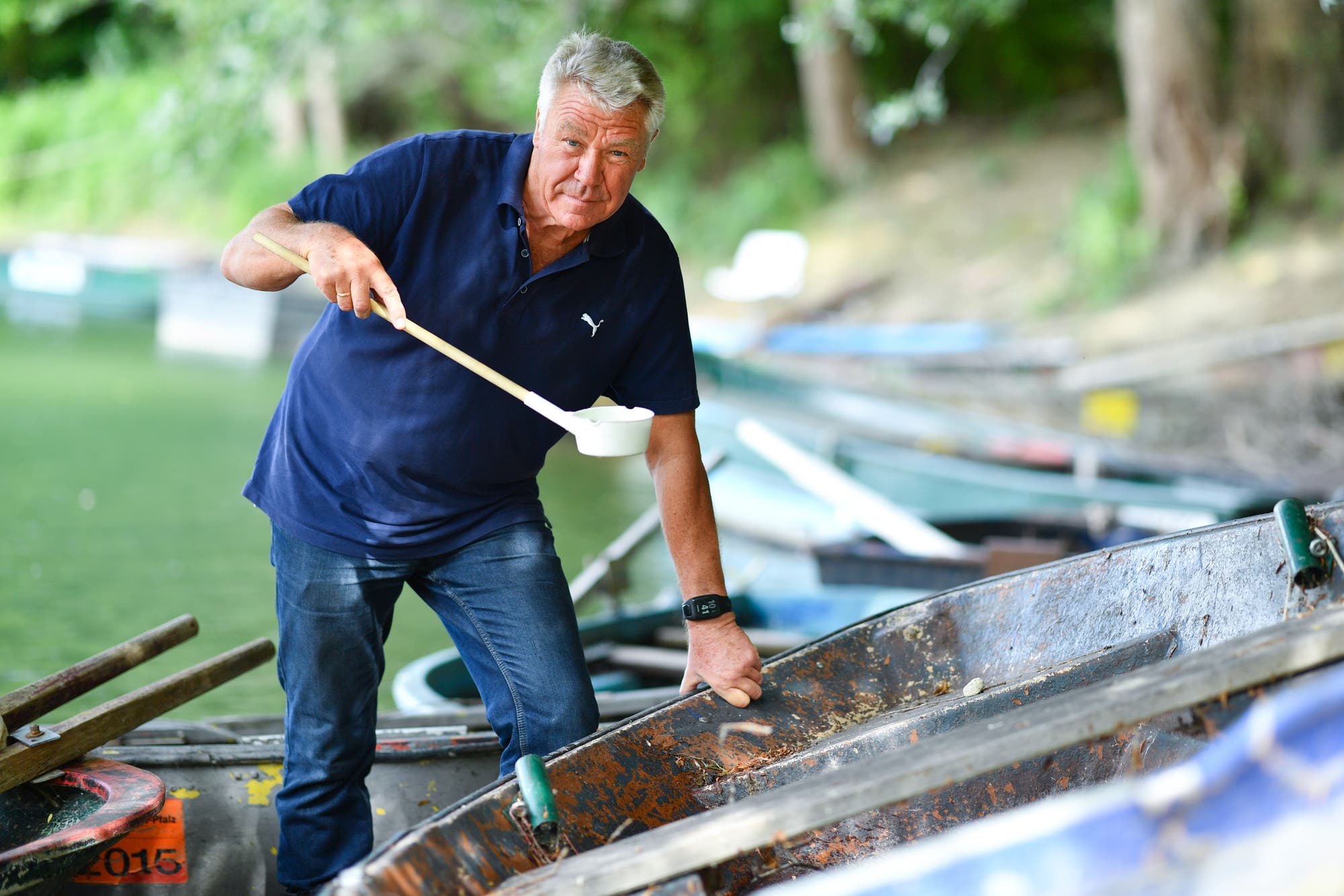 Das Bild zeigt den Wissenschaftler Norbert Becker, der in Speyer (Rheinland-Pfalz) am Ufer des Altrheins steht und ein Probengefäß mit Wasser in der Hand hält. Er hat blaue Kleidung an. Im Hintergrund sieht man Boote am Ufer sowie flussbegleitende Pflanzen.