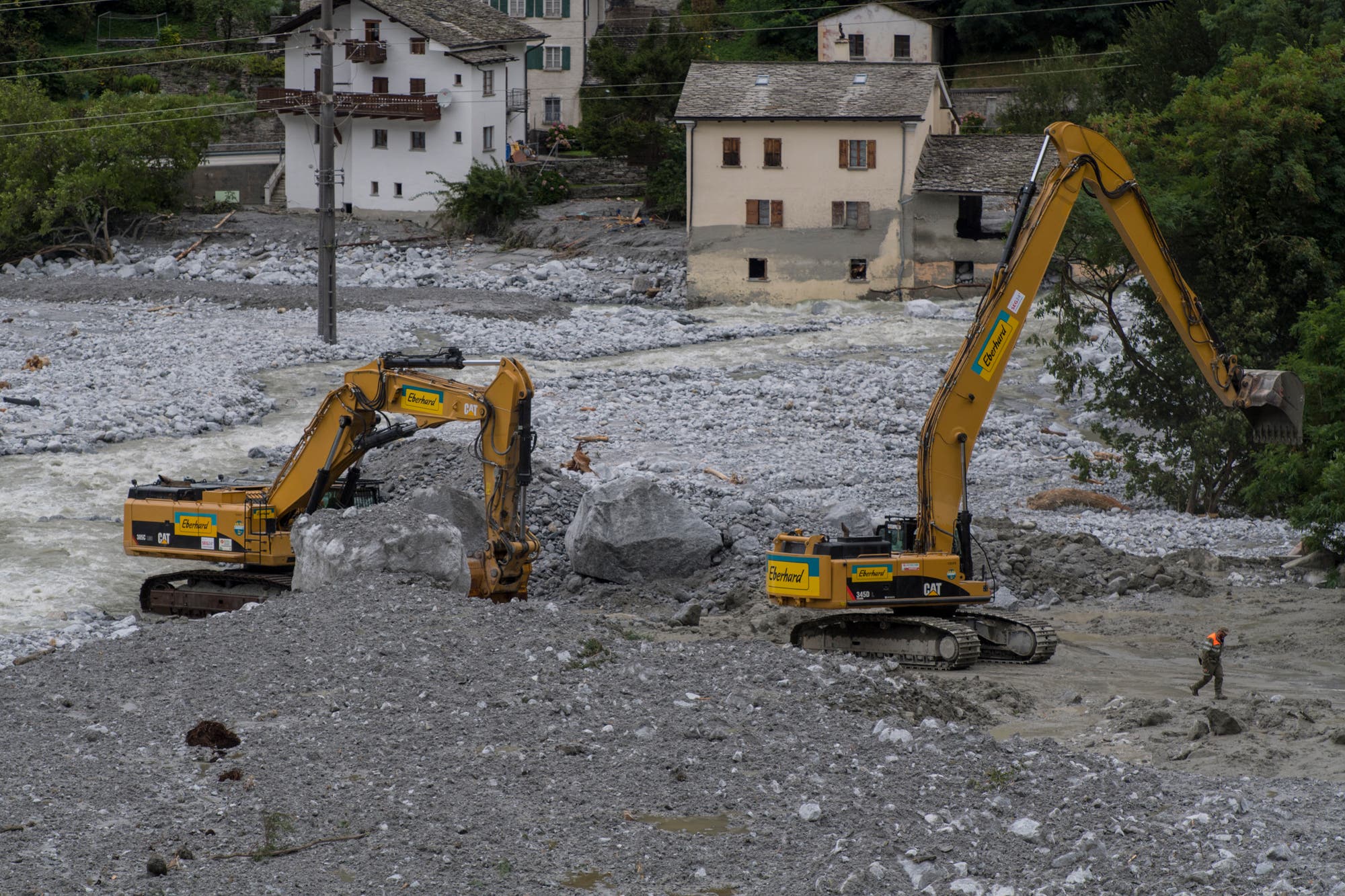 Zwei gelbe Bagger der Marke Eberhard arbeiten in einer steinigen Flusslandschaft, umgeben von Geröll und großen Felsen. Im Hintergrund sind mehrere zweistöckige Häuser mit Schindeldach zu sehen. Ein Arbeiter mit orangefarbenem Helm steht rechts neben den Baggern. Die Szene zeigt eine Baustelle in einer alpinen Umgebung.