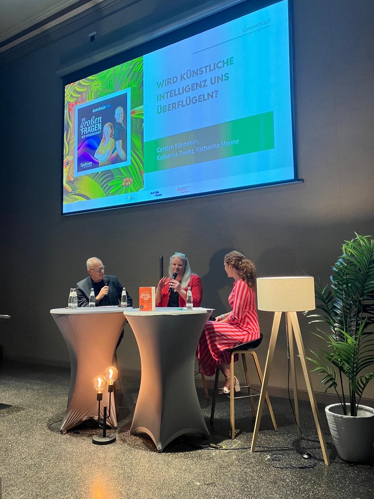 Eine Podiumsdiskussion mit drei Personen, die an einem Tisch sitzen. Im Hintergrund ist eine große Leinwand mit der Aufschrift „Wird künstliche Intelligenz uns überflügeln?“ zu sehen. Auf dem Tisch stehen Wasserflaschen und ein orangefarbenes Buch. Eine Stehlampe und eine Pflanze befinden sich rechts im Bild. Die Szene findet in einem modernen, gut beleuchteten Raum statt.