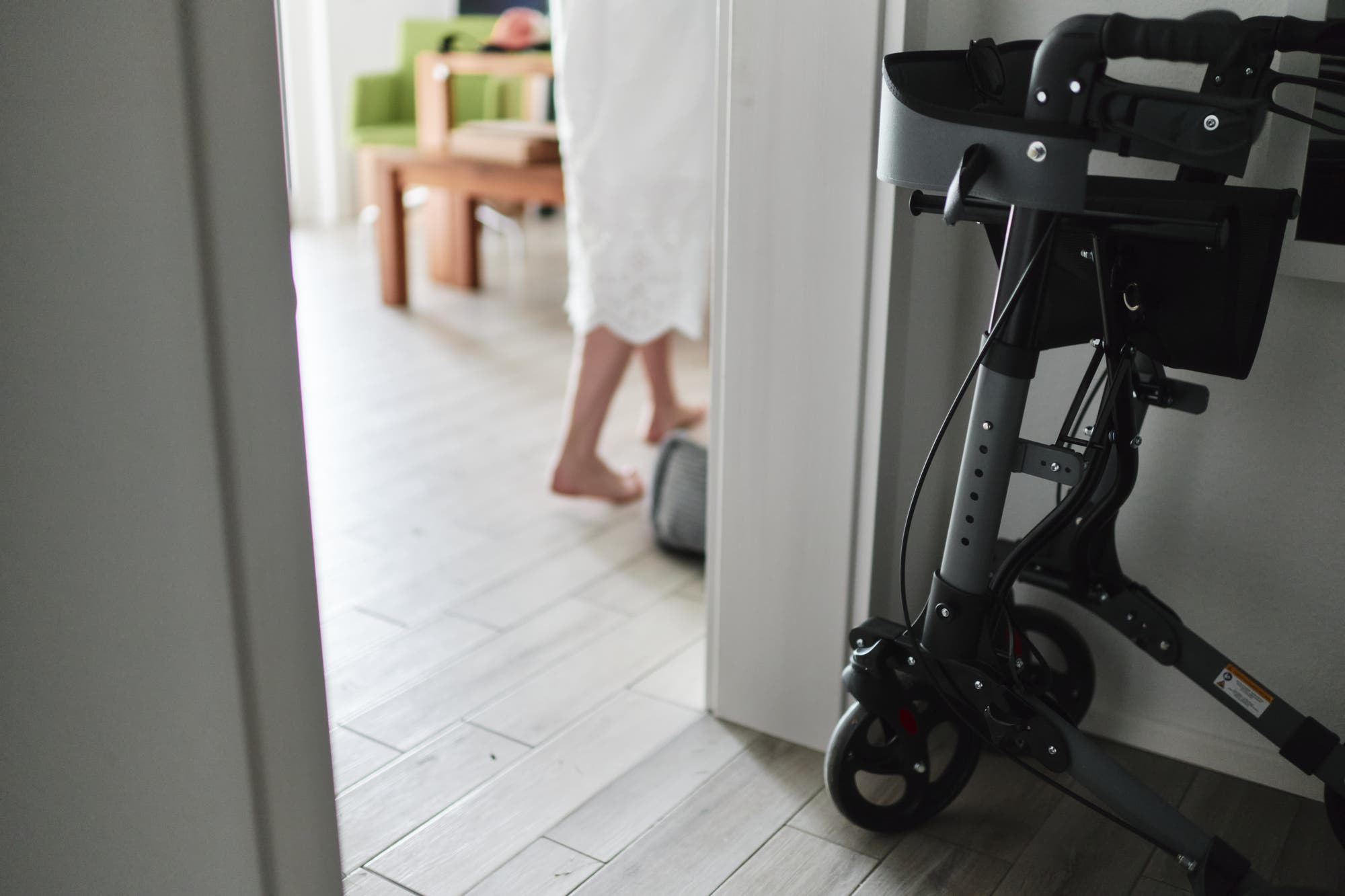 Eine Person in einem weißen Kleid läuft barfuß in einen Raum. Im Vordergrund ist ein Rollator zu sehen. 