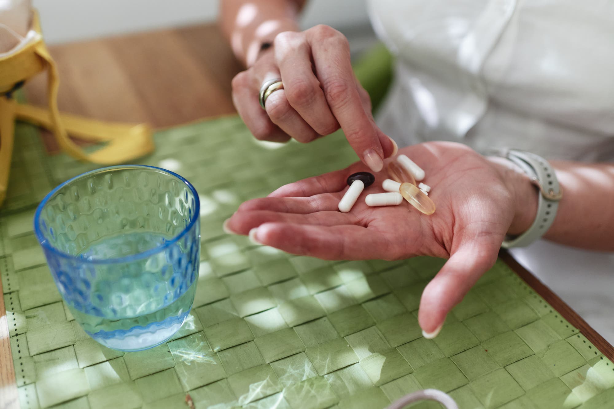 Natalie Grams hält mehrere Kapseln und Tabletten in der Hand, während sie mit der anderen Hand eine davon auswählt. Sie trägt Ringe und eine Uhr. Auf dem Tisch liegt eine grüne Tischdecke, daneben steht ein blaues Glas mit Wasser. 