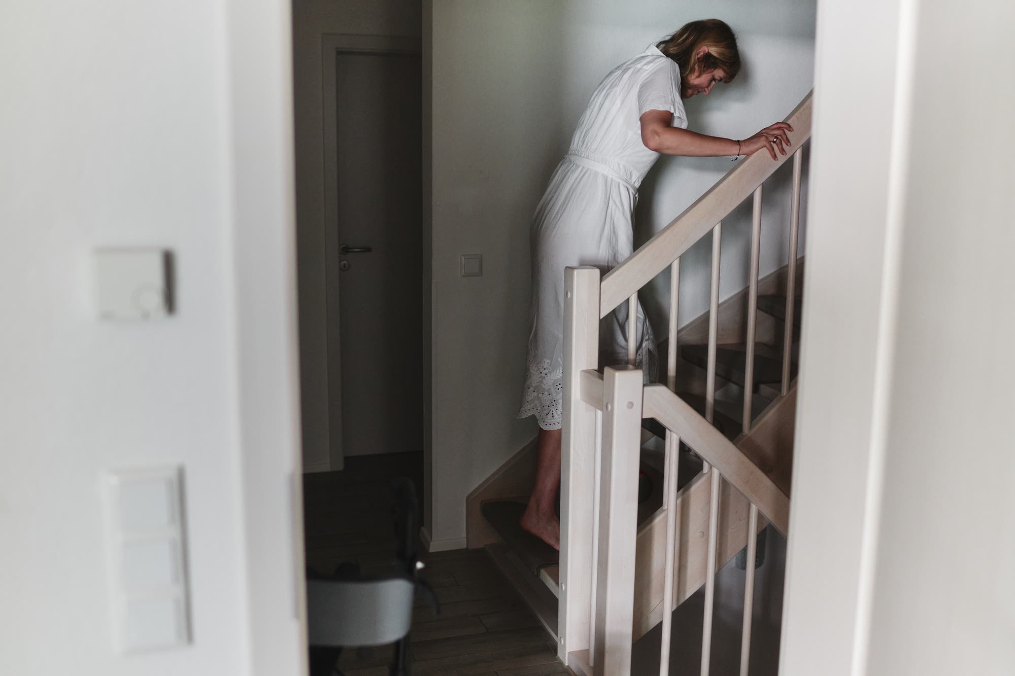 Die an Long Covid und ME/CFS erkrankte Ärztin und Autorin Natalie Grams steigt in einem weißen Kleid eine Holztreppe im Flur hinauf. Sie hält sich am Geländer fest und bewegt sich vorsichtig nach oben. Im Vordergrund ist ein Teil eines Rollstuhls sichtbar.