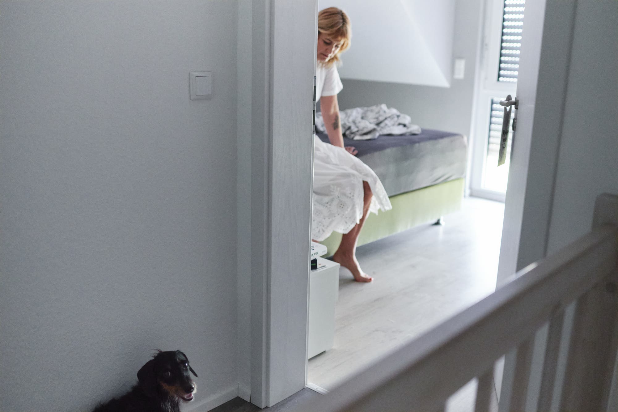 Eine Frau sitzt auf einem Bett in einem hellen Raum, der durch eine offene Tür sichtbar ist. Sie trägt ein weißes Kleid und schaut nach unten. Im Vordergrund sitzt ein kleiner schwarzer Hund auf dem Boden im Flur. Der Raum ist minimalistisch eingerichtet, mit einem Fenster, das Tageslicht hereinlässt.