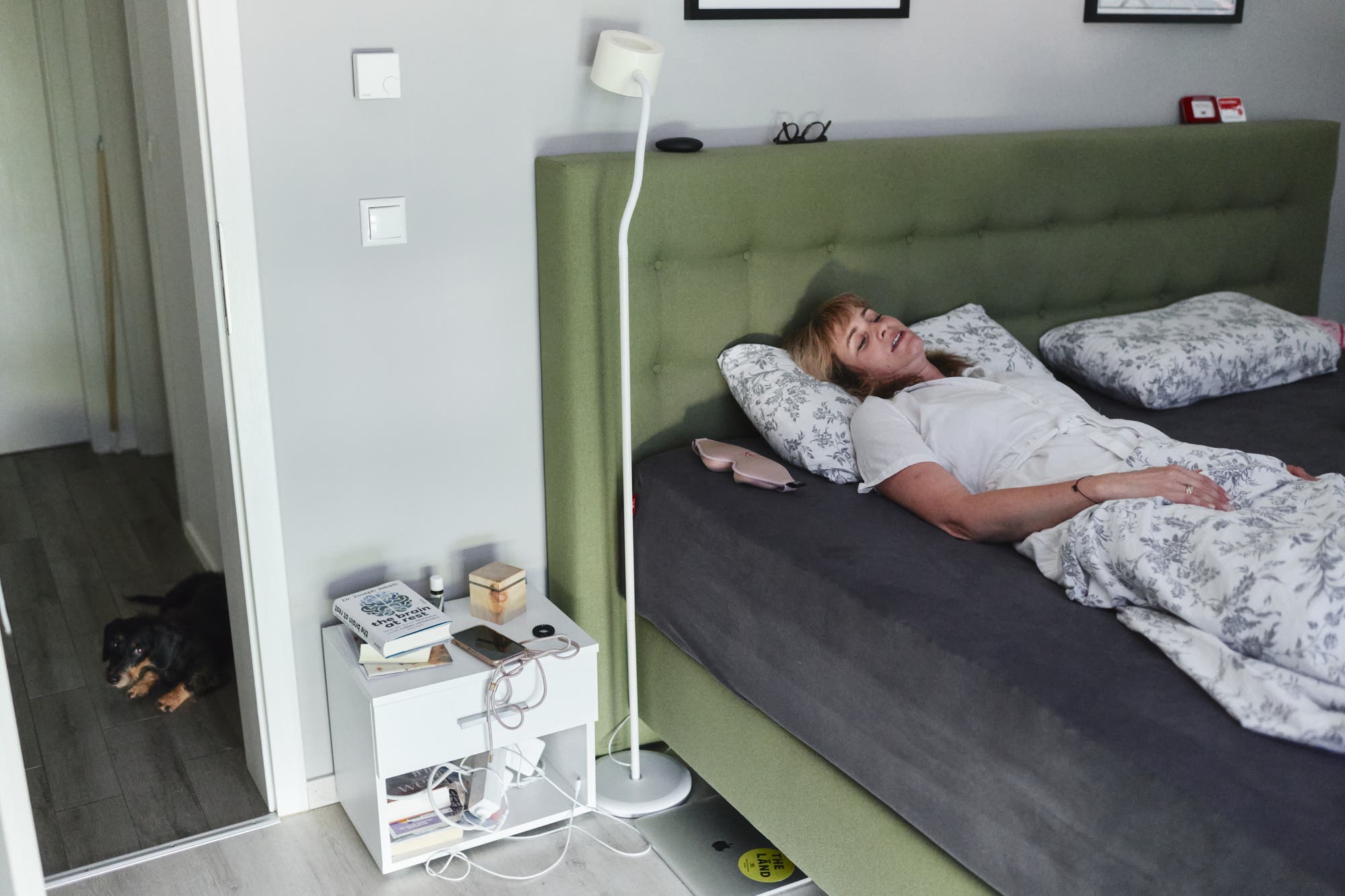 Die an ME/CFS erkrankte Natalie Grams liegt auf einem Bett mit grünem Kopfteil, umgeben von Kissen mit Blumenmuster. Neben dem Bett steht ein Nachttisch mit Büchern, einer Brille und einem Smartphone. Eine Stehlampe steht daneben. Im Hintergrund ist ein Hund im Türrahmen zu sehen. Der Raum wirkt gemütlich und modern eingerichtet.