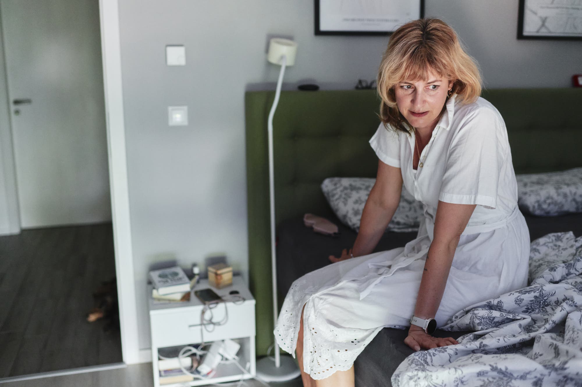 Natalie Grams sitzt in einem weißen Kleid auf einem Bett mit einem grünen Kopfteil und gemusterten Bettdecken. Neben dem Bett steht ein kleiner Nachttisch mit Büchern, einem Smartphone und anderen Gegenständen. Die an ME/CFS erkrankte Autorin und Ärztin wirkt nachdenklich und schaut zur Seite.