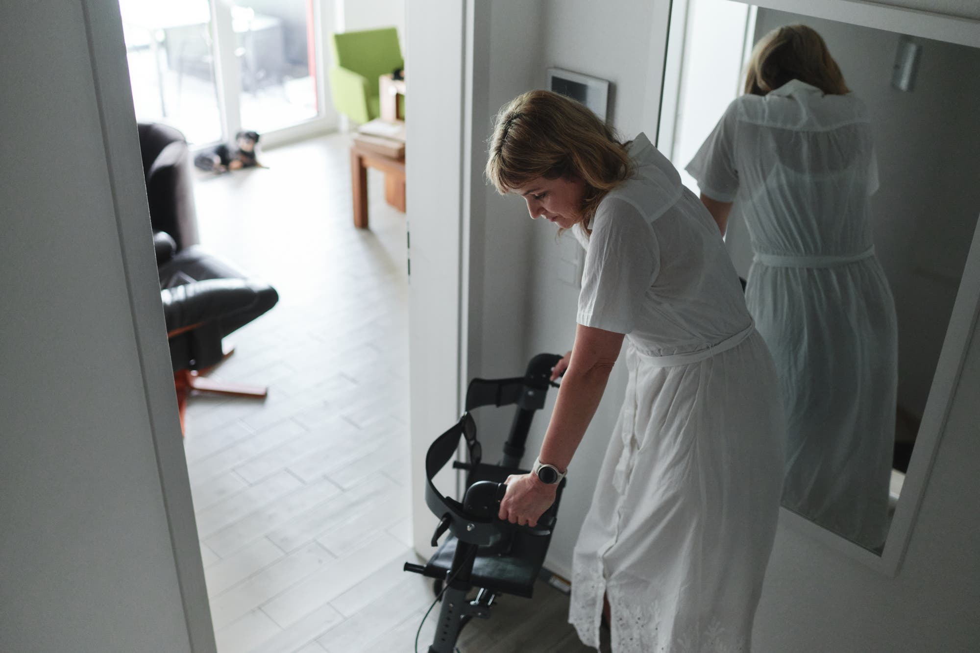 Die Autorin Natalie Grams trägt ein weißes Kleid und hält sich an einen Rollator fest. Im Hintergrund sind ein schwarzer Sessel, ein grüner Stuhl und ein Tisch zu sehen. Ein Hund liegt auf dem Boden. 