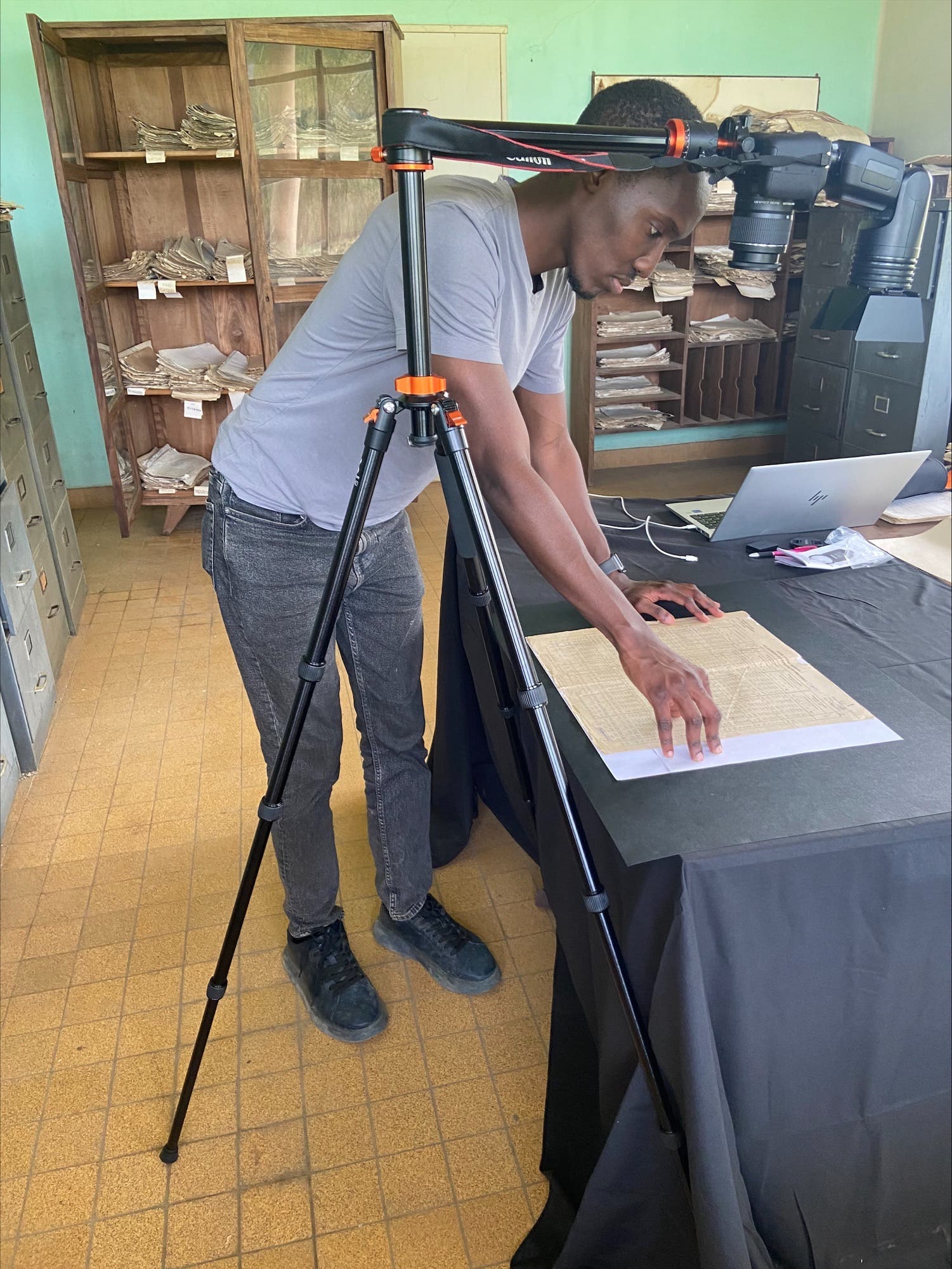 Ein Wissenschaftler fotografiert historische Dokumente in einem Raum mit Regalen voller Akten. Er verwendet eine Kamera auf einem Stativ, um die Dokumente auf einem Tisch zu erfassen. Ein Laptop steht daneben, möglicherweise zur Datenverarbeitung. Der Raum hat grüne Wände und einen Holzboden.