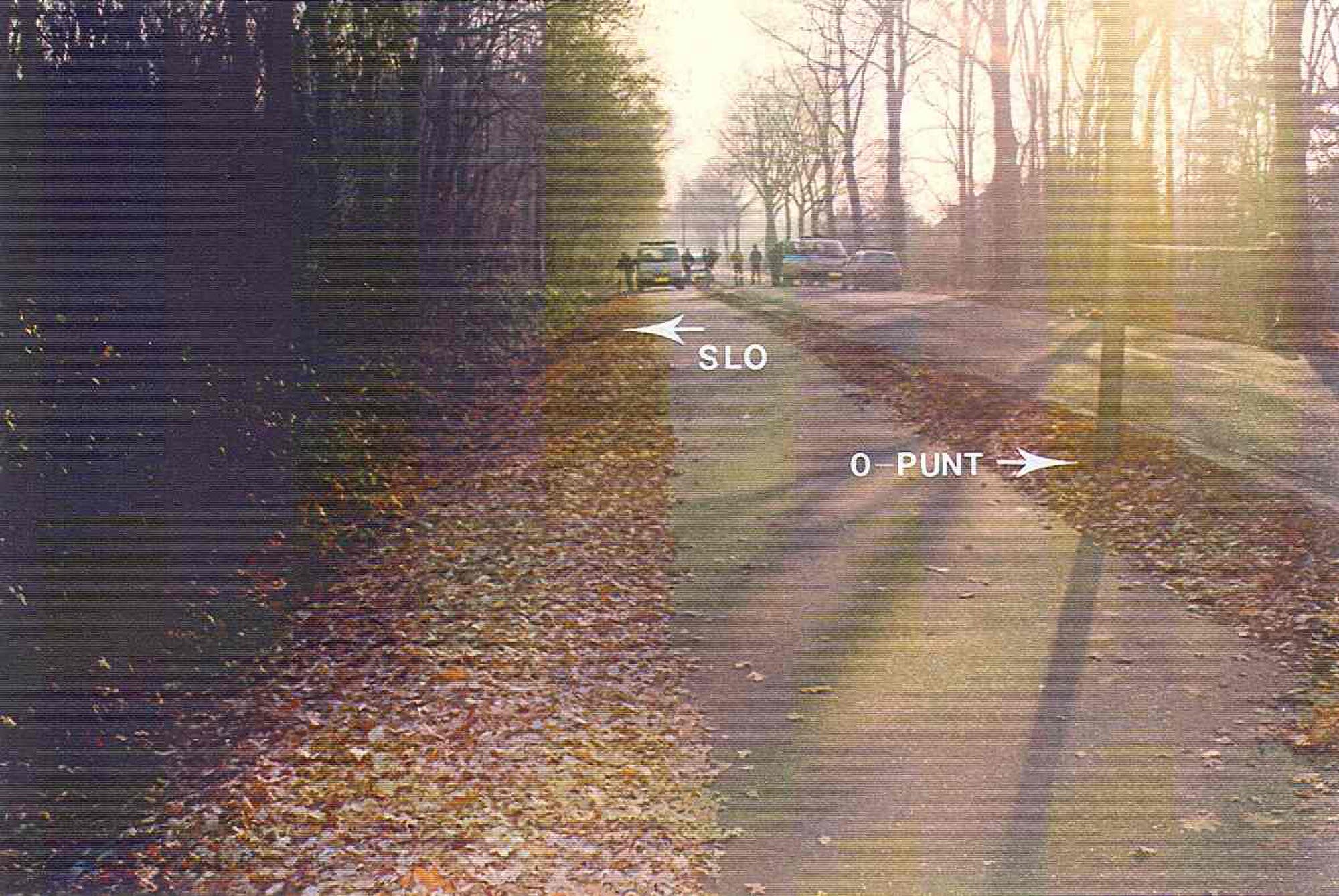 Ein herbstlicher Weg mit Laub bedeckt, der durch einen Wald führt. Links ist dichter Wald, rechts eine Straße mit Autos. Zwei Pfeile zeigen auf den Weg: einer mit der Aufschrift "SLO" und der andere "0-PUNT". Im Hintergrund sind ein Polizeiwagen sowie Menschen zu sehen, die auf der Straße gehen.