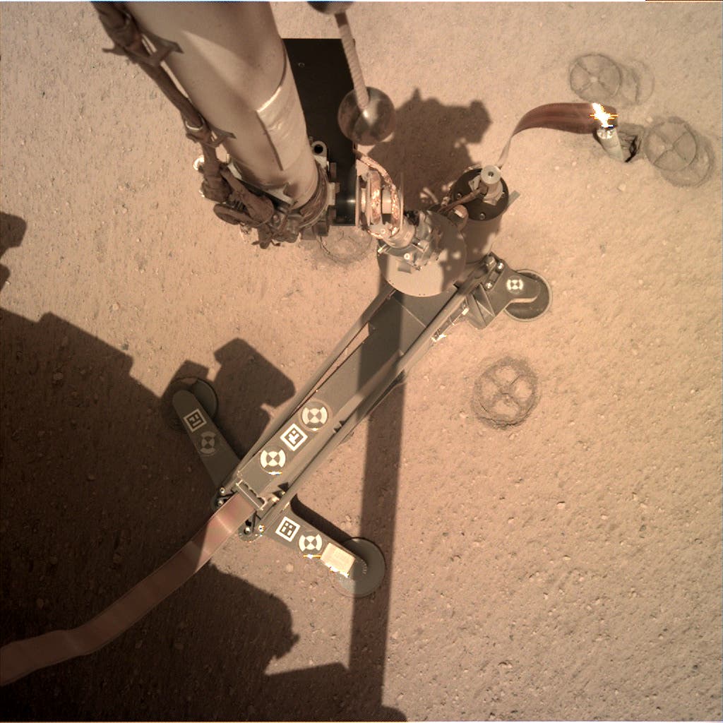 Ein Roboterarm der Marssonde InSight berührt die sandige Oberfläche des Mars. Der Arm ist mit verschiedenen technischen Komponenten ausgestattet, darunter Kabel und Sensoren. Auf dem Boden sind kreisförmige Abdrücke zu sehen. Die Szene ist gut beleuchtet und zeigt die detaillierte Struktur des Marsbodens.