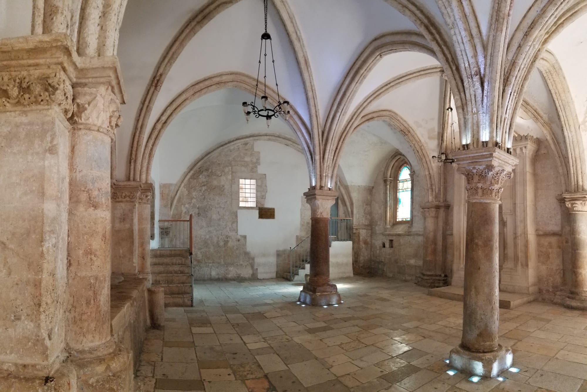Ein historischer Raum mit gotischen Gewölbedecken und steinernen Säulen. Die Wände sind aus hellem Stein, und es gibt bunte Glasfenster, die sanftes Licht hereinlassen. Ein Kronleuchter hängt von der Decke. Der Boden besteht aus großen, rechteckigen Steinplatten. Im Hintergrund sind Treppen zu sehen, die zu einer erhöhten Plattform führen.