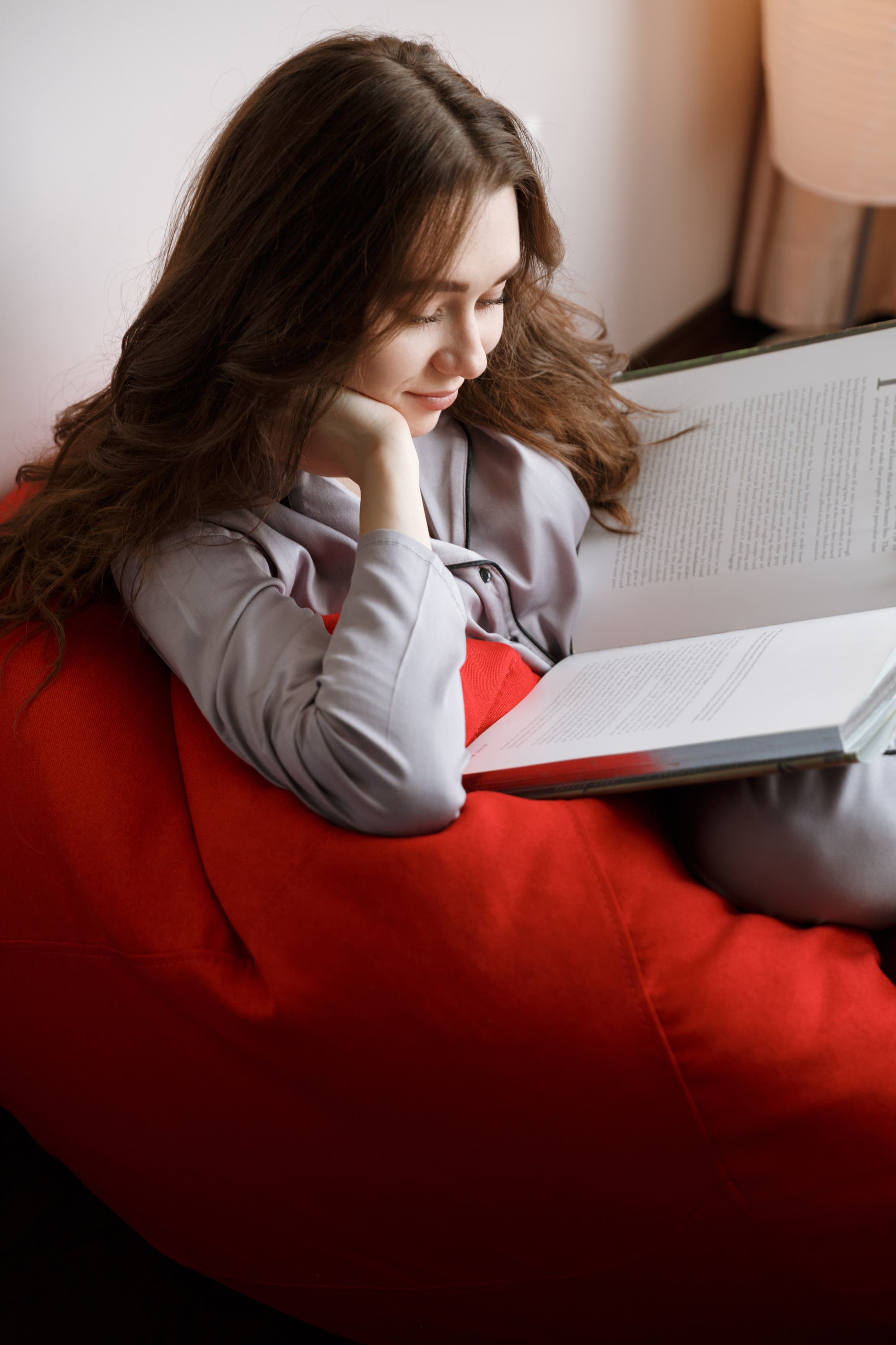 Eine junge brünette Frau sitzt mit aufgestütztem Kopf auf einem roten Sofa und liest ein Buch.
