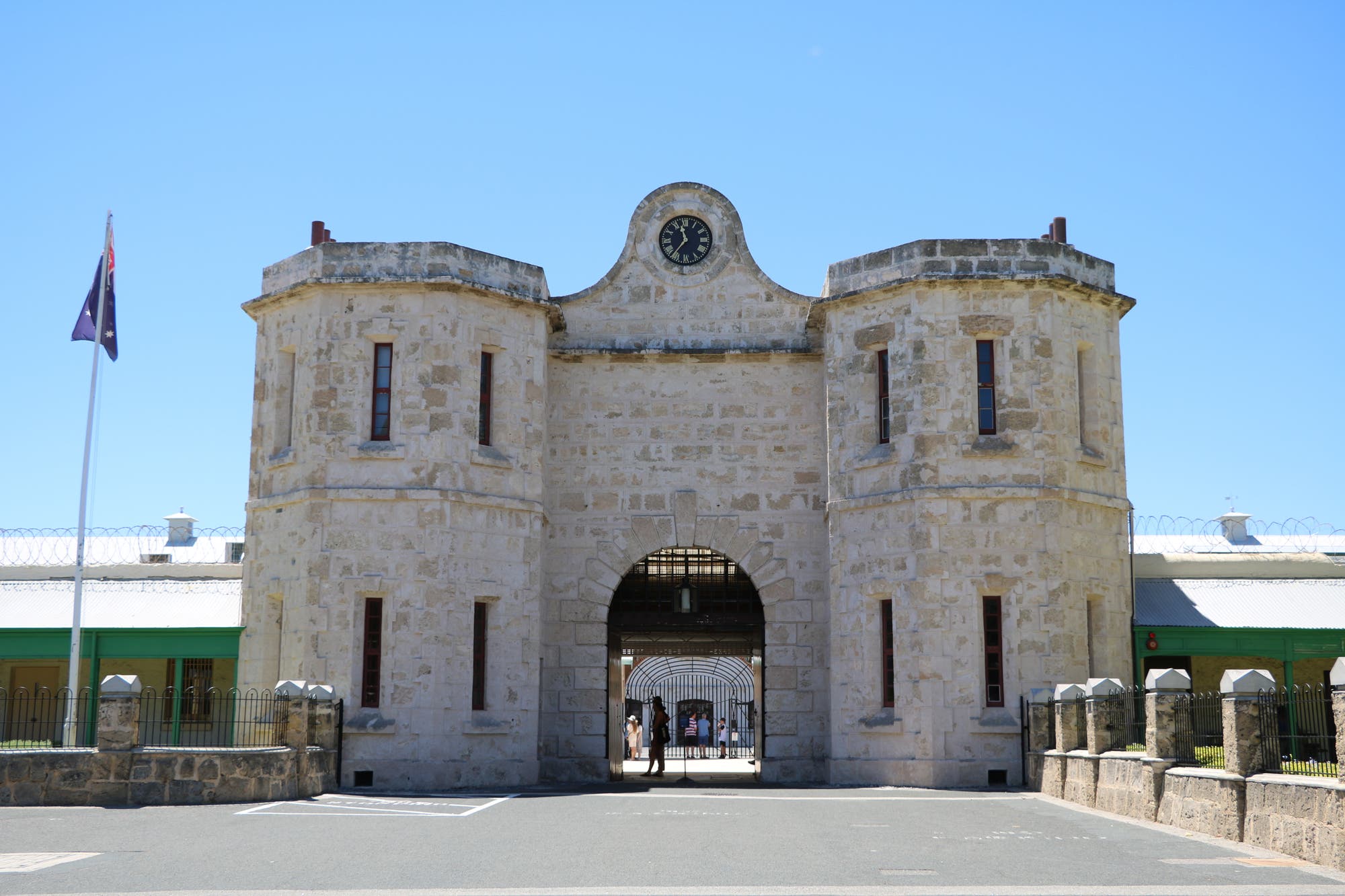 Historisches Gefängnisgebäude aus hellem Stein mit zwei runden Türmen und einer zentralen Uhr über dem Eingang. Eine australische Flagge weht links im Bild. Menschen gehen durch das Tor. Der Himmel ist klar und blau.