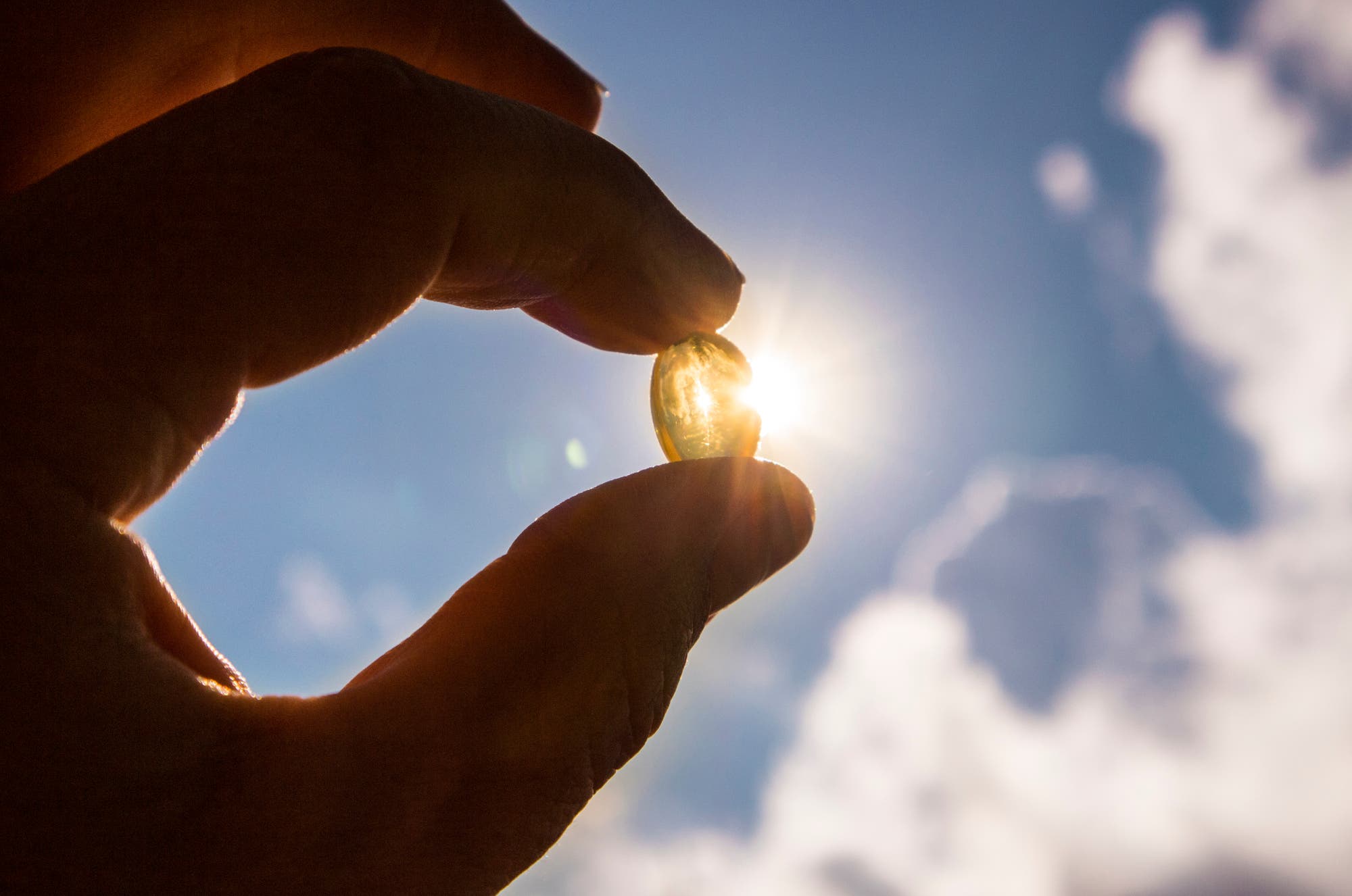 Eine Hand hält eine gelblich-durchsichtige Kapsel gegen den Himmel, und zwar so, dass die Sonne durch diese scheint. 