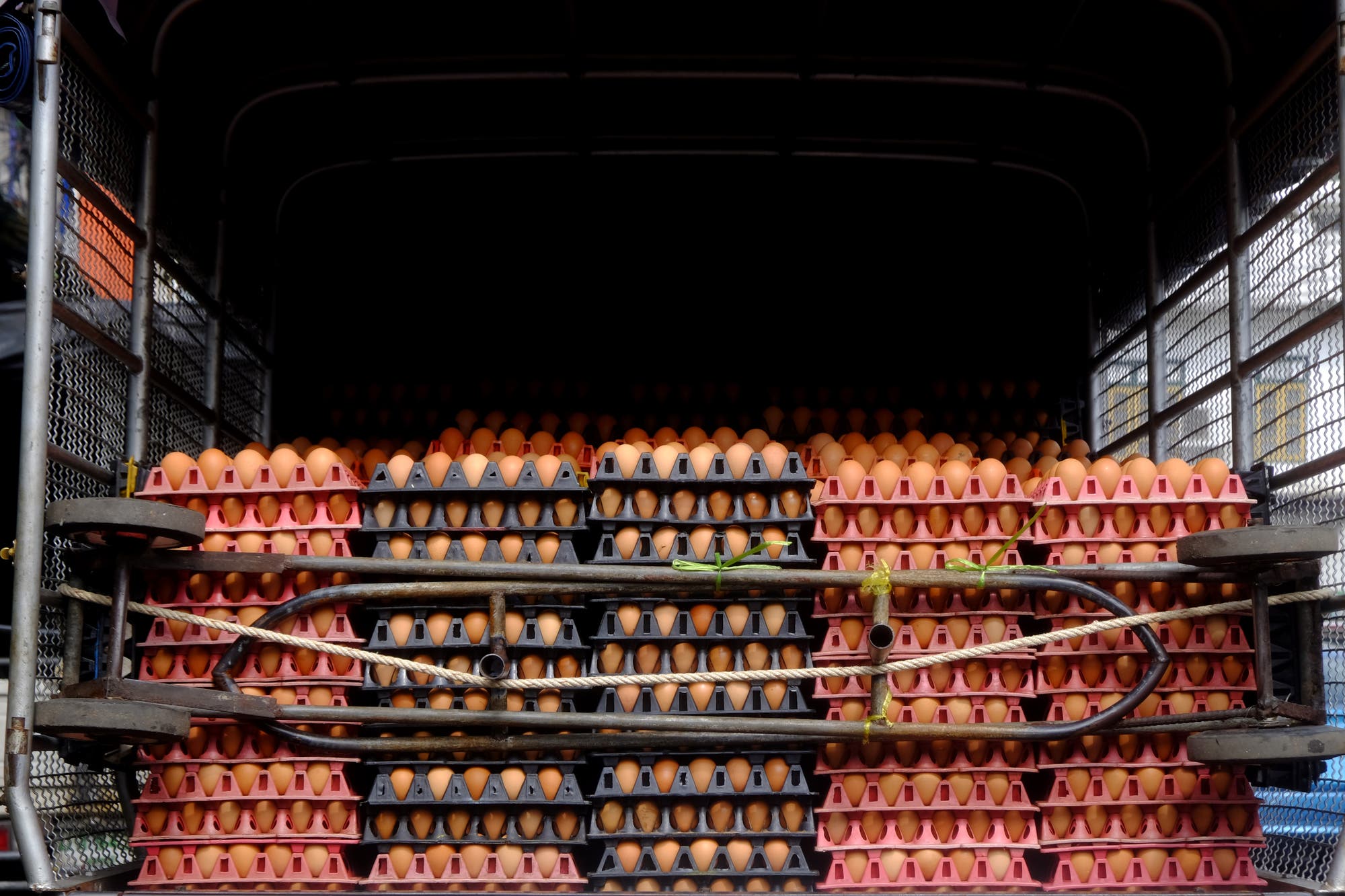 Ein Lkw mit offenen Türen, beladen mit mehreren Stapeln von Eiern in bunten Kunststoffträgern. Die Eier sind in schwarzen und rosa Trays gestapelt und durch Seile gesichert. Der Innenraum des Lkw ist dunkel, während die Eier im Vordergrund gut sichtbar sind. Es gibt keine Personen im Bild.