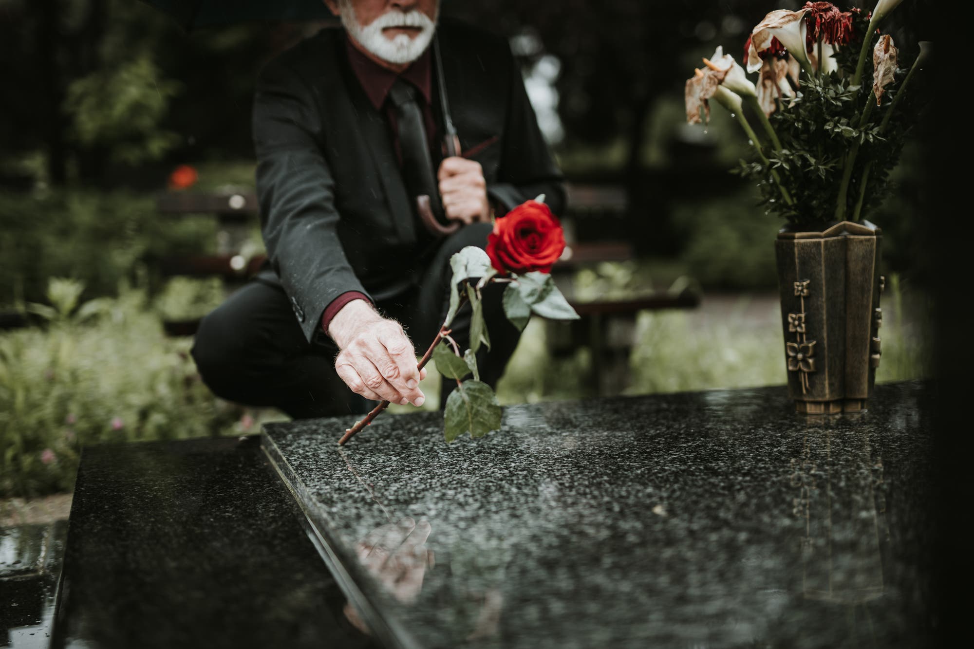 Ein älterer Mann mit weißem Bart, in einem dunklen Anzug und mit Regenschirm, legt eine rote Rose auf einen glänzenden Grabstein. Neben dem Grab steht eine Vase mit verwelkten Blumen.