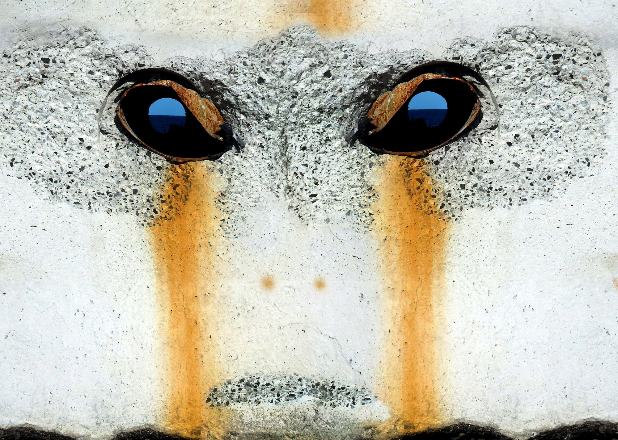 Eine Nahaufnahme einer Betonoberfläche mit zwei dunklen, runden Öffnungen, die wie Augen wirken. Die Oberfläche zeigt unregelmäßige, graue und orangefarbene Muster, die an ein Gesicht erinnern. Die Textur des Betons ist rau und körnig.