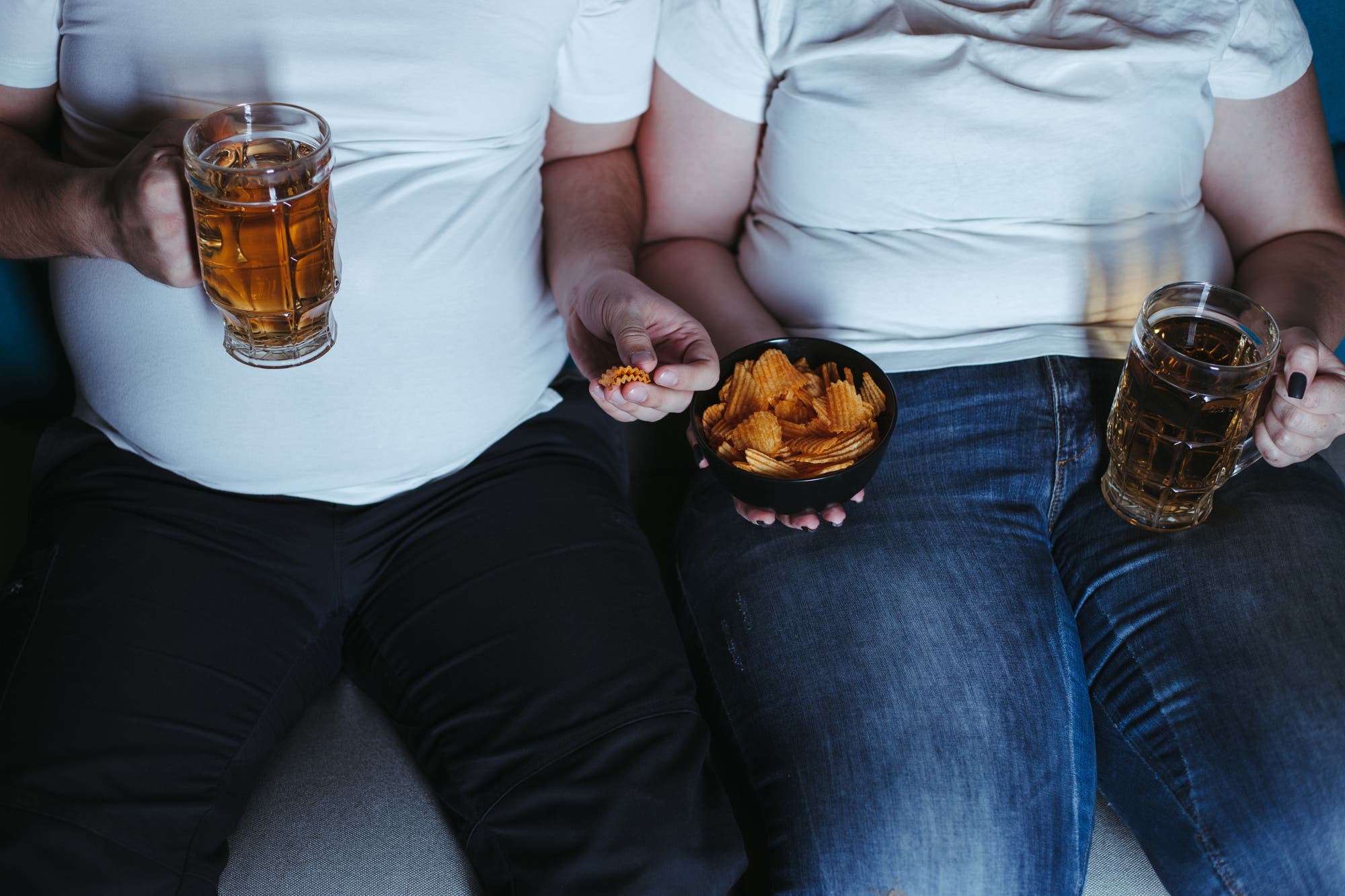 Zwei übergewichtige Personen in weißen T-Shirts sitzen nebeneinander auf einem Sofa. Beide halten ein Glas Bier in der Hand. Die Person links hält zusätzlich eine kleine Schale mit Chips, während die Person rechts eine Chipstüte hält.
