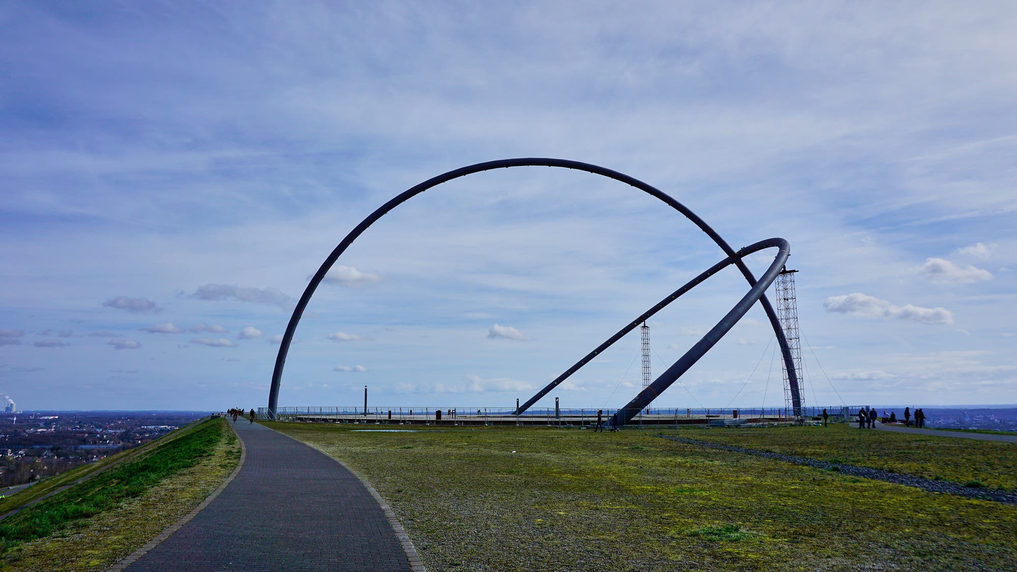 Man blickt seitlich auf das Horizontobservatorium auf der Halde Hoheward. Zwei mächtige Bögen mit einem Radius von mehr als 45 Metern markieren Meridian und Äquator der Erd- und der Himmelskugel. Der Äquatorbogen wird an zwei Stellen abgestützt. Die Silhouetten einiger Menschen sind klein im Hintergrund erkennbar.
