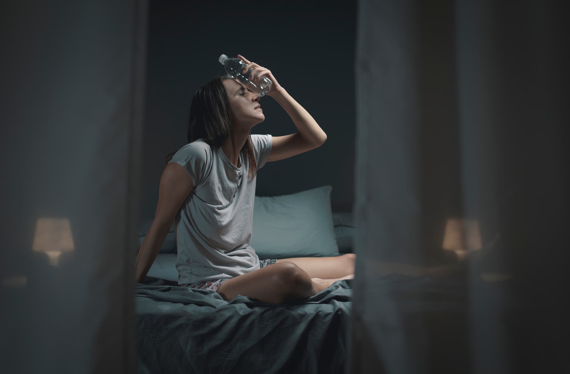 Eine Frau sitzt aufrecht im Bett und hält sich eine Flasche Wasser an den Kopf