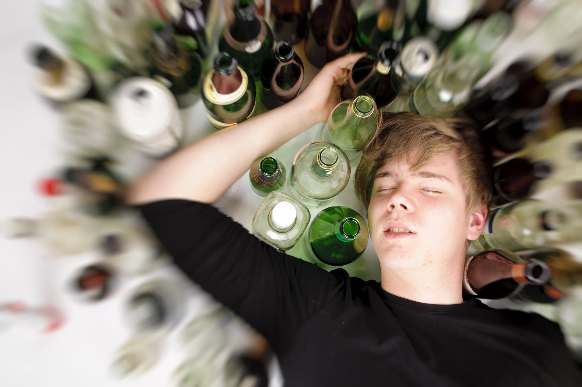 Ein männlicher Jugendlicher liegt im Rausch schlafend neben vielen leeren Flaschen auf dem Boden.