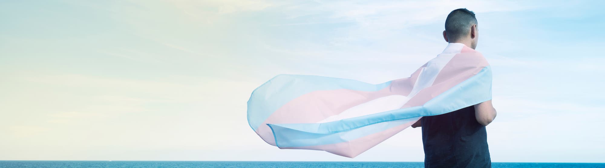 Eine Person steht mit dem Rücken zur Kamera und blickt auf das Meer. Sie trägt eine transgender Flagge in den Farben Blau, Pink und Weiß um die Schultern, die im Wind weht.