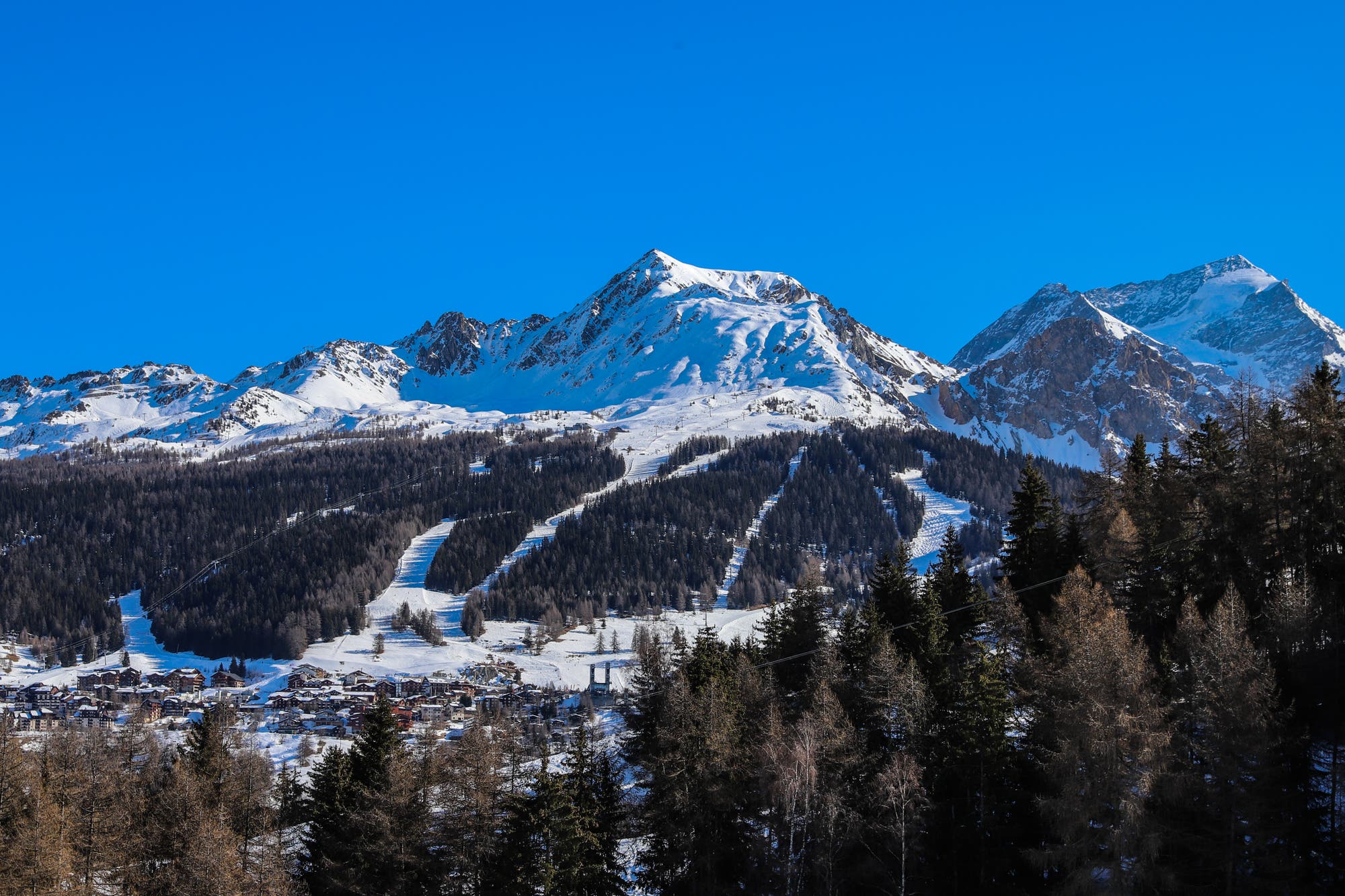 Blick auf einen schneebedeckten Berggipfel unter blauen Himmel. Darunter erstreckt sich ein kleines Dorf mit einigen Dutzend Häusern im Chalet-Stil.