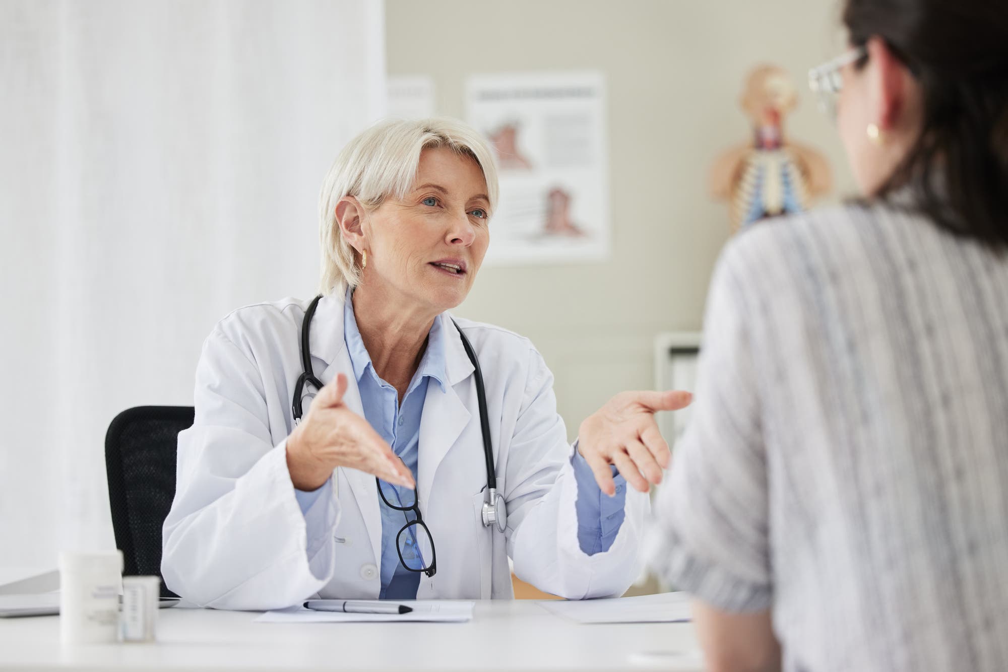 Eine Ärztin mit grauem Haar und Stethoskop spricht mit einer Patientin in einem Behandlungszimmer. Die Ärztin gestikuliert, während sie der Patientin zuhört. Im Hintergrund sind medizinische Diagramme sichtbar.