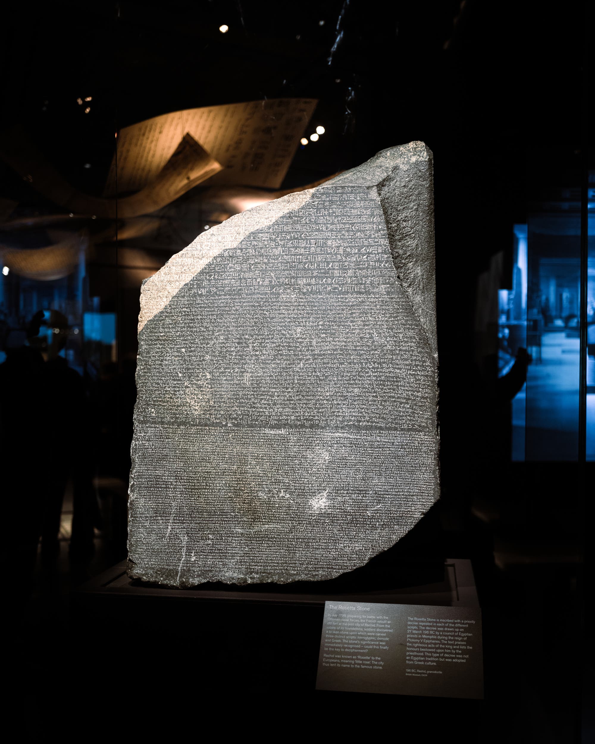 Eine große, unvollständige Steintafel mit eingravierten Texten in drei verschiedenen Schriften, bekannt als der Stein von Rosetta, ausgestellt im British Museum. Der Stein ist von einem dunklen Raum umgeben, mit schwachem Licht, das seine Oberfläche beleuchtet. Im Hintergrund sind unscharfe Silhouetten von Menschen zu erkennen, die die Ausstellung betrachten. Eine Informationstafel vor dem Stein bietet Details zur Geschichte und Bedeutung des Artefakts. Der Stein von Rosetta ist ein Schlüssel zur Entzifferung ägyptischer Hieroglyphen.