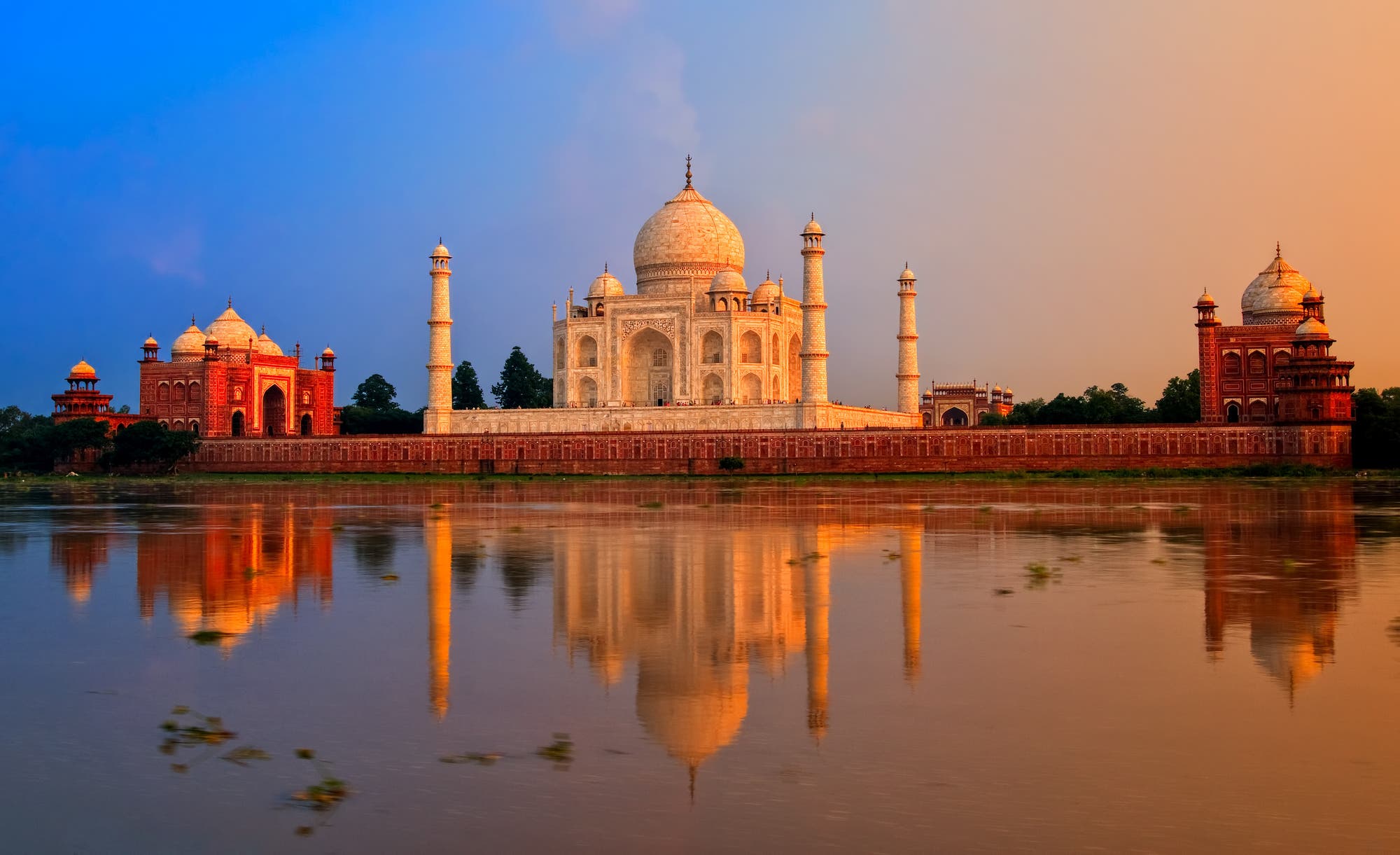Ein beeindruckendes Foto des Taj Mahal bei Sonnenuntergang, das sich im ruhigen Wasser eines Flusses spiegelt. Der Himmel zeigt einen sanften Übergang von Blau zu Orange, während das weiße Marmor-Mausoleum von zwei roten Sandstein-Gebäuden flankiert wird. Die Szene strahlt Ruhe und Erhabenheit aus.
