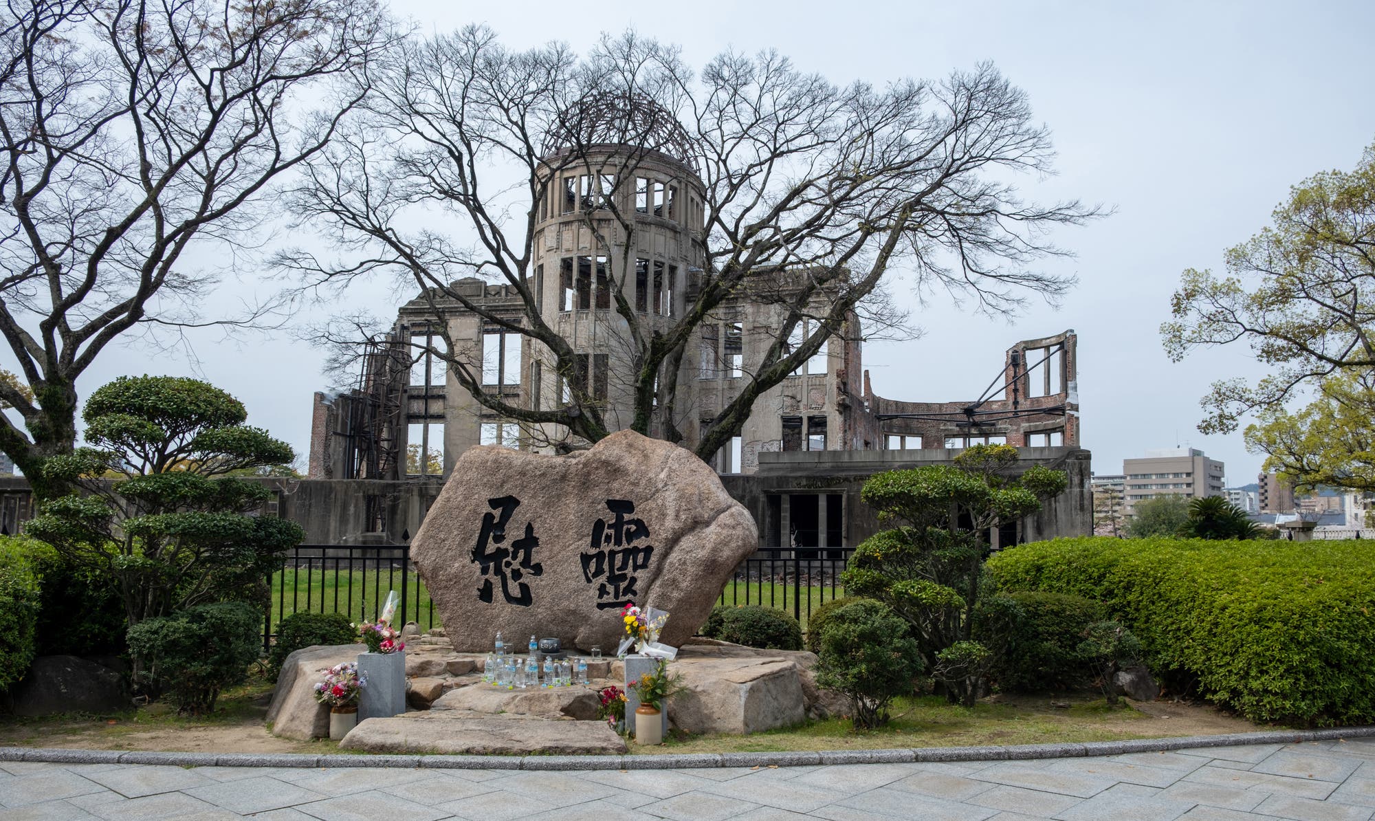 Eine Fotografie des Atombomben-Doms in Hiroshima, Japan. Im Vordergrund steht ein Gedenkstein mit japanischen Schriftzeichen, umgeben von Blumen und Kerzen. Der Dom, eine Ruine mit einer markanten Kuppel, ist von kahlen Bäumen und gepflegtem Grün umgeben. Der Himmel ist bewölkt.