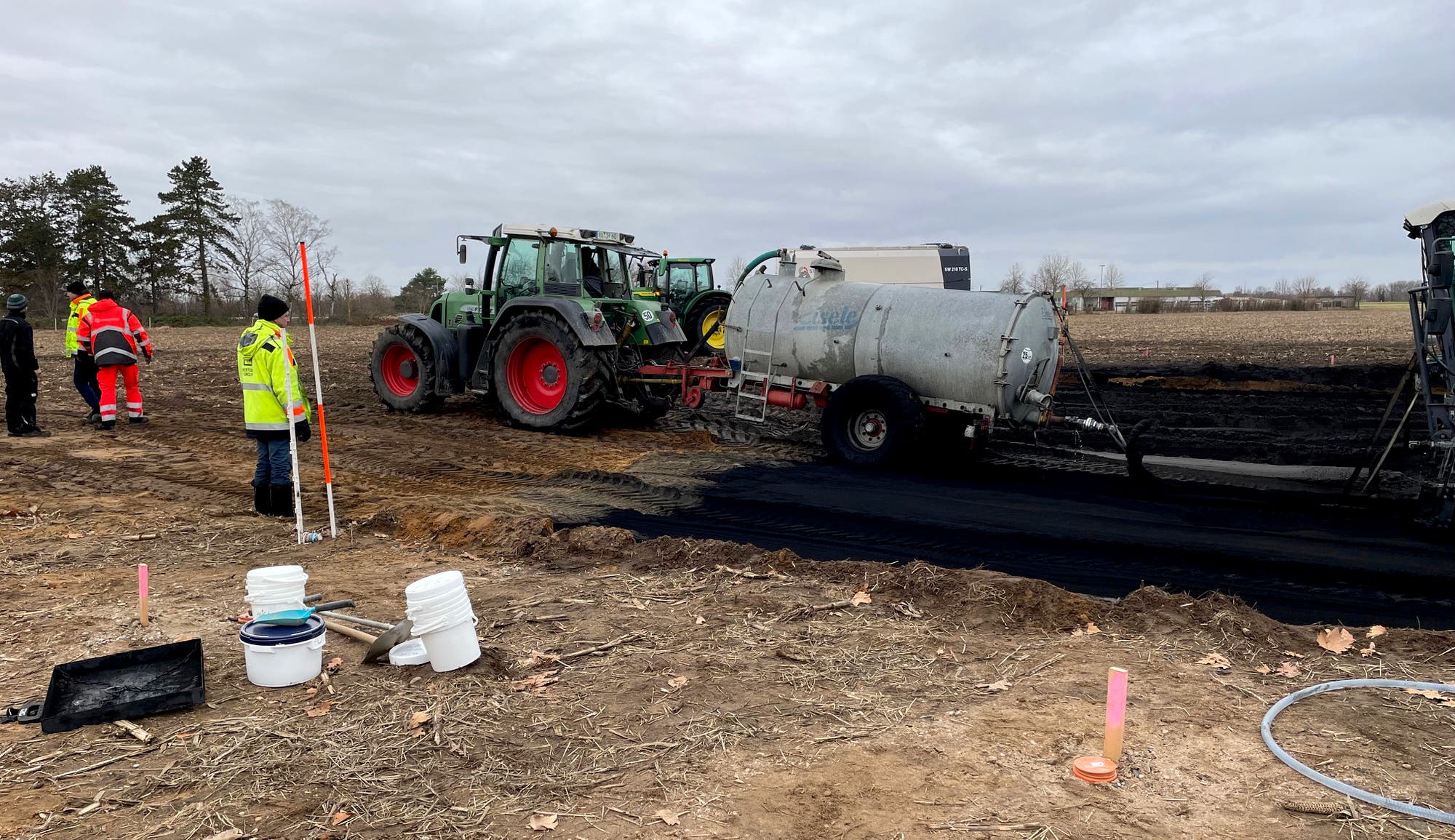 Ein Traktor zieht einen Tankanhänger über ein Feld, während Arbeiter in Warnkleidung die Szene beobachten. Der Boden ist teilweise schwarz bedeckt, möglicherweise durch eine Substanz aus dem Tankanhänger. Im Vordergrund stehen Eimer und Werkzeuge auf dem Boden. Der Himmel ist bewölkt, und im Hintergrund sind Bäume und ein Gebäude zu sehen.