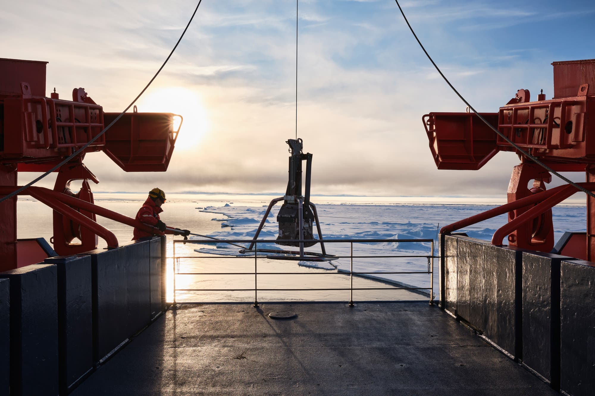 ein Foto zeigt einen Kran, der eine Probe an Bord der Polarstern hievt. im Hintergrund eisbedecktes Meer und Sonne hinter Wolken