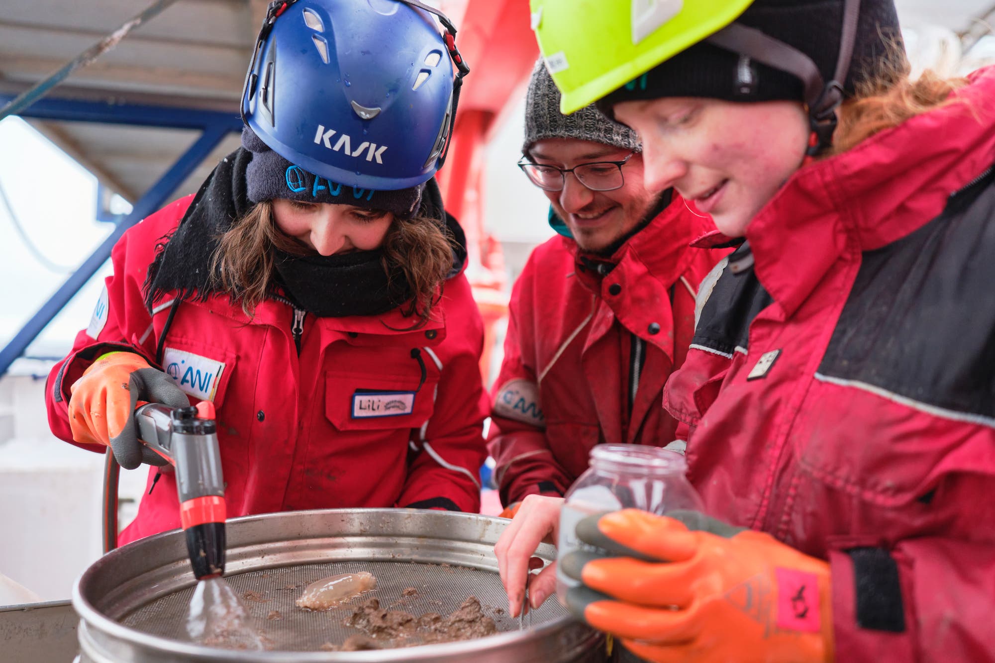 Ein Foto zeigt drei Wissenschaftlerinnen in roten Polaranzügen, die mit einer Handbrause schlammige Proben über einem Metallsieb abbrausen.