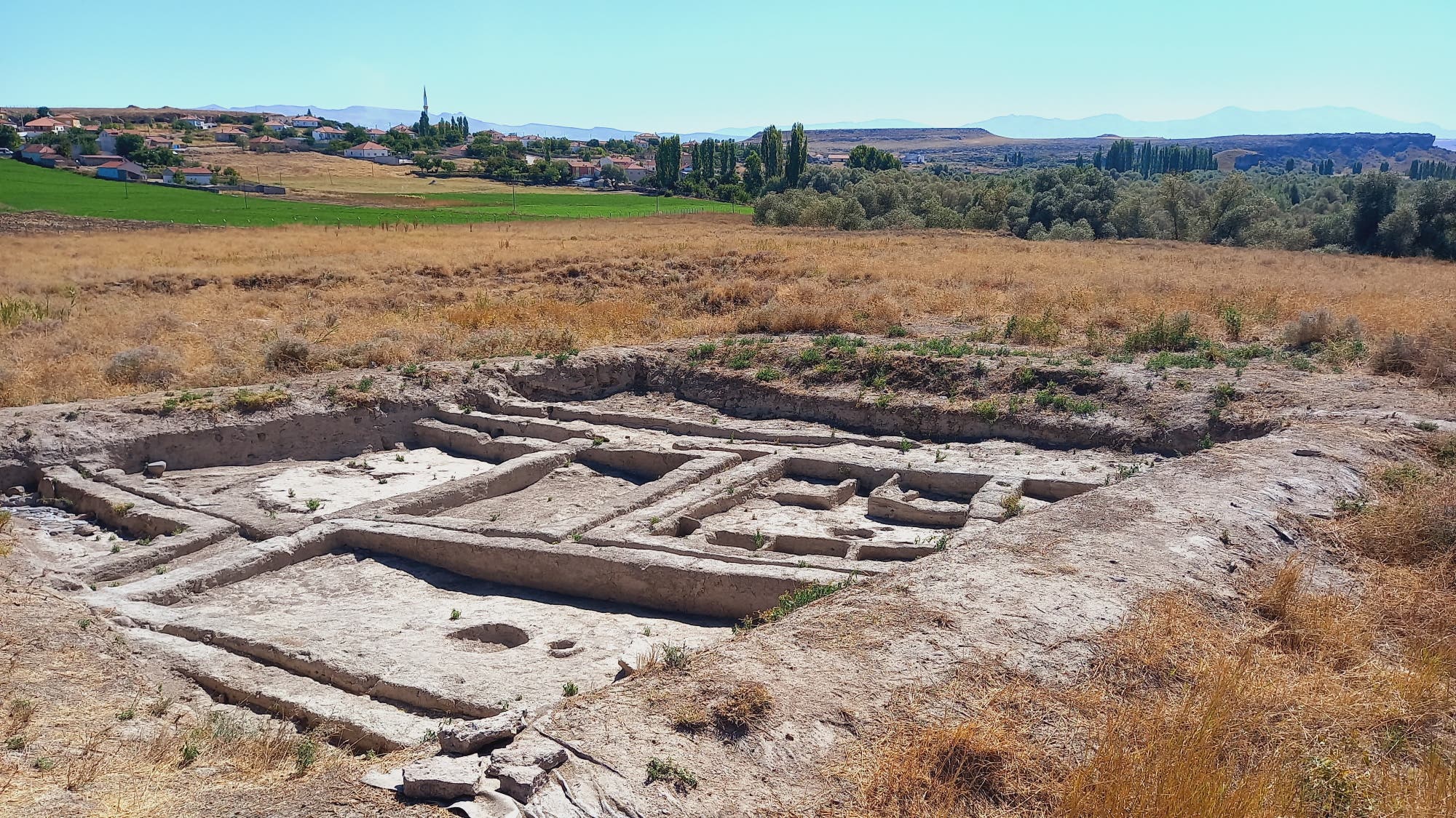 Archäologische Ausgrabungsstätte von Aşıklı Höyük in einer ländlichen Landschaft. Im Vordergrund sind die frei gelegten Überreste von neolithischen Strukturen zu sehen. Im Hintergrund erstrecken sich Felder und eine kleine Siedlung mit Häusern und Bäumen. Die Szene ist von Hügeln und einem klaren Himmel umgeben.