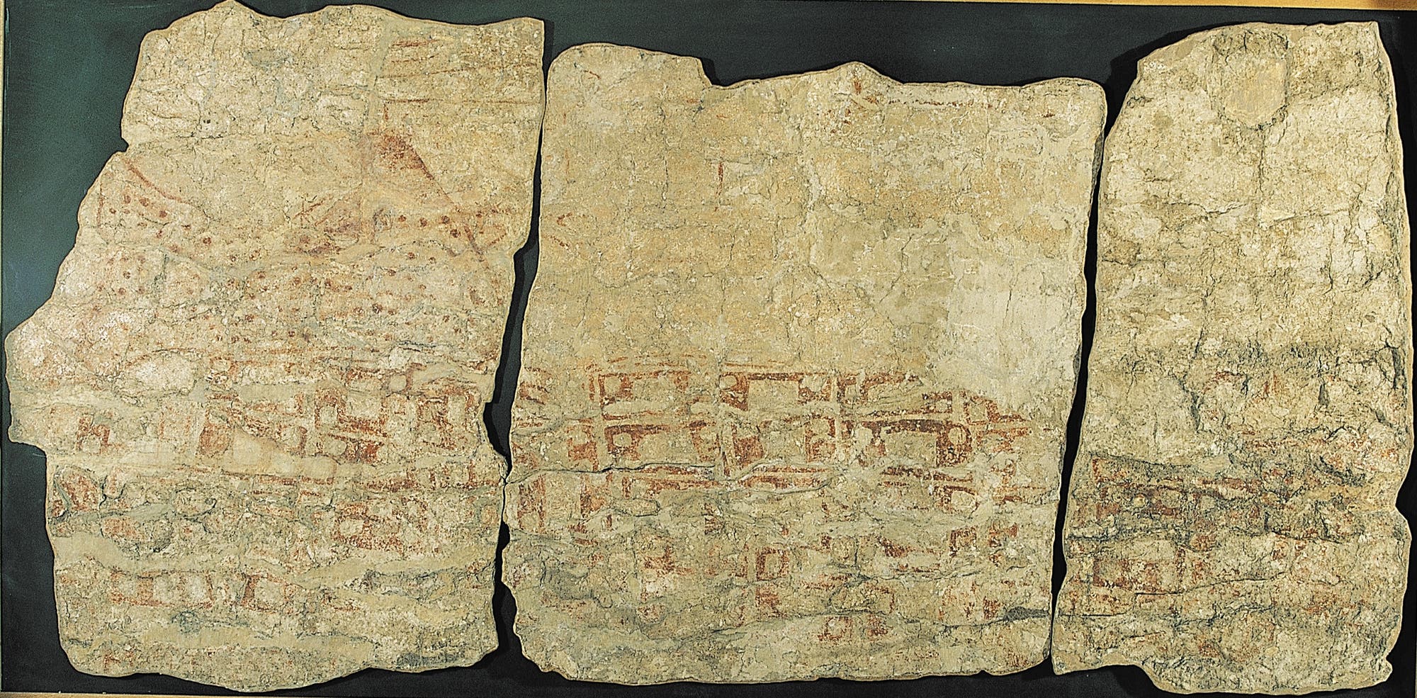 Drei Wandmalereifragmente aus Çatalhöyük sind in einem Holzrahmen ausgestellt. Die Fragmente zeigen verblasste rote und braune Rechtecke und einen Vulkanberg mit doppelter Spitze auf einem beigen Hintergrund. 
