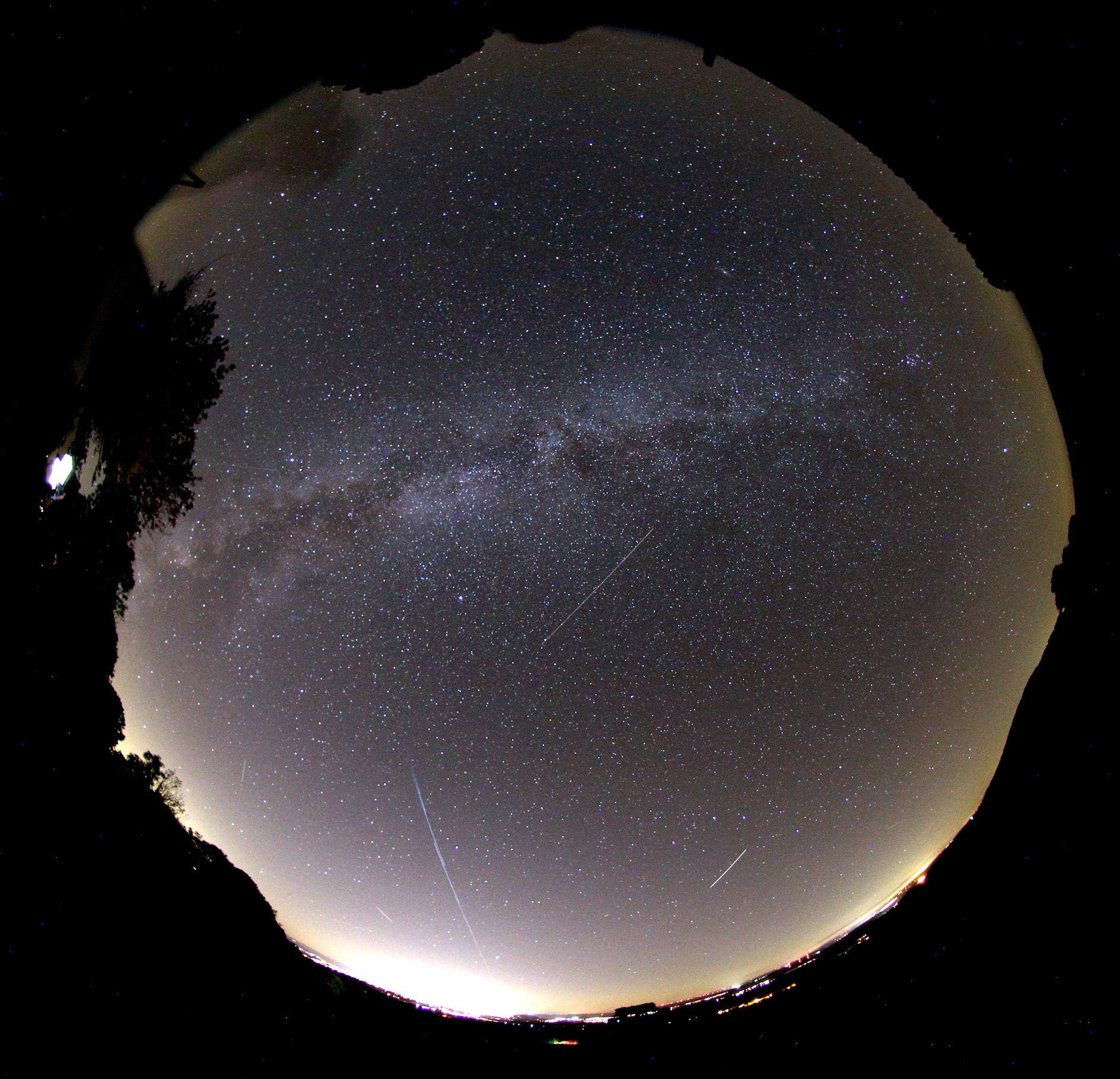 Die Aufnahme mit einem Fischaugenobjektiv zeigt große Teile des dunklen Himmels mit punktförmigen Sternen und Strichspuren von Satelliten
