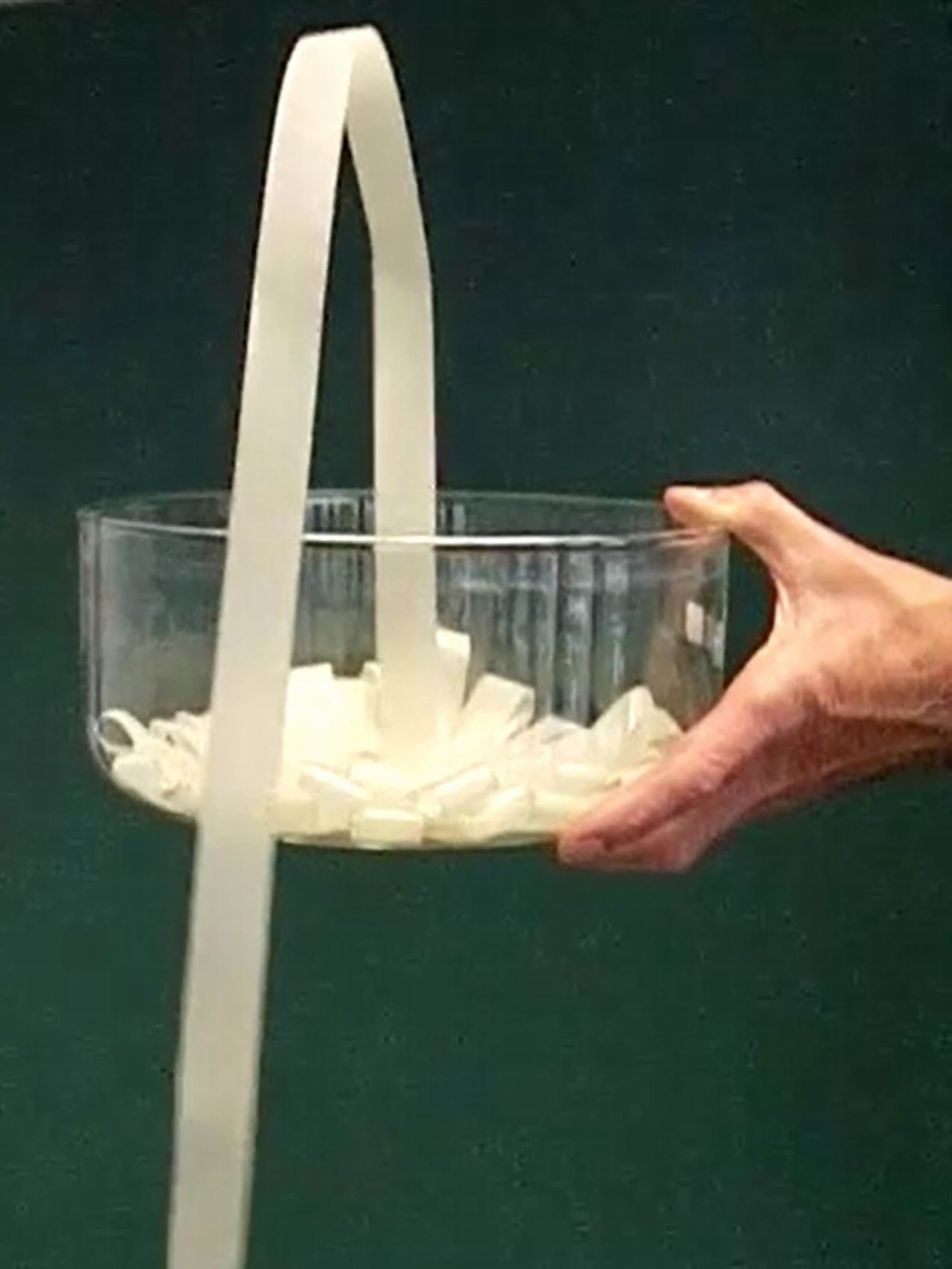Dieses Einzelbild aus einem Video zeigt, wie ein zusammengelegtes Band aus einer hochgehaltenen Glasschale springt, über deren Rand hinaus zu Boden.