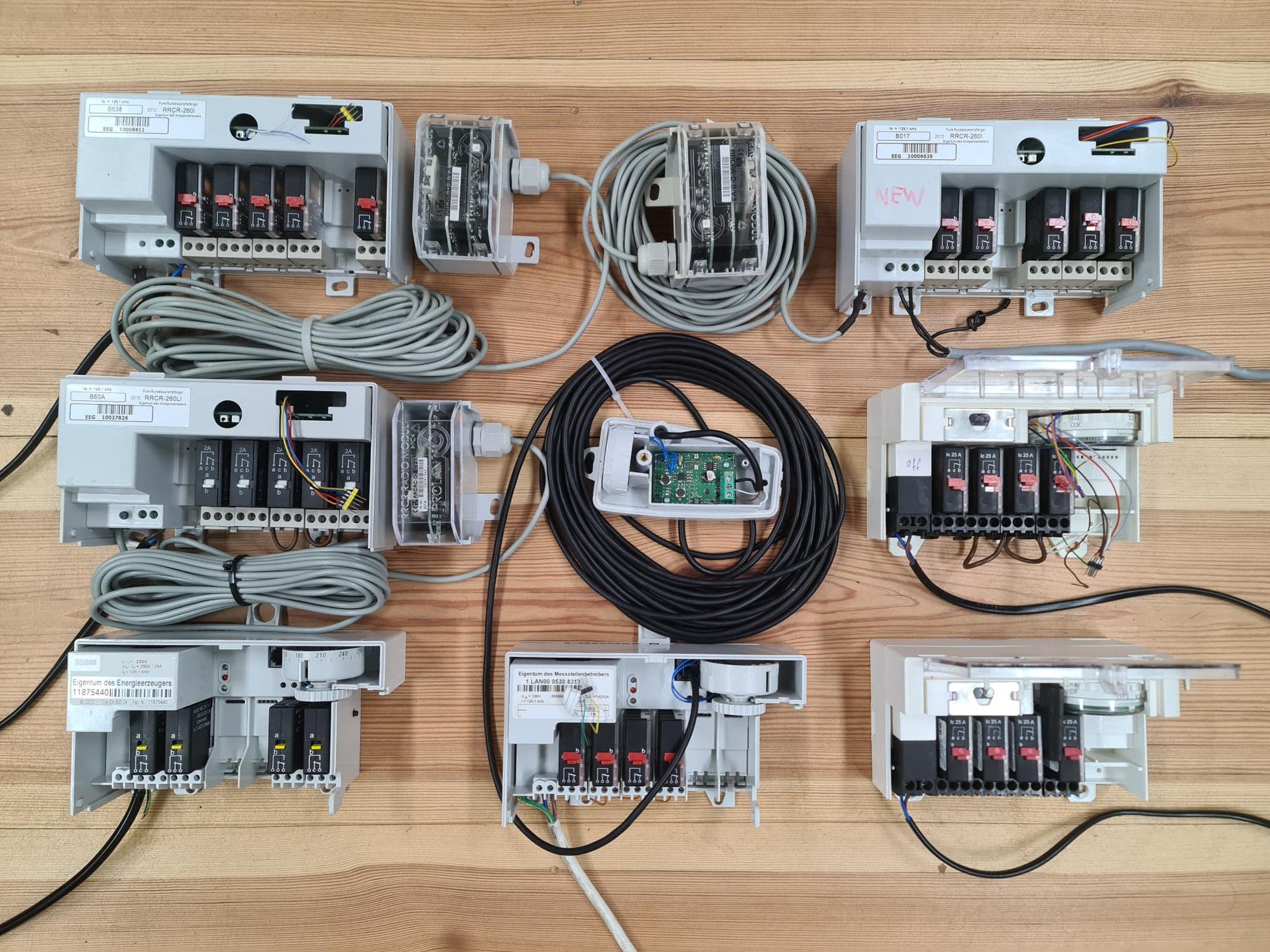 Eine Sammlung von elektrischen Schaltkästen und Kabeln auf einem Holztisch. Die Schaltkästen sind geöffnet und zeigen verschiedene Sicherungen und Verkabelungen. In der Mitte befindet sich ein Gerät mit einer grünen Leiterplatte und einem langen schwarzen Kabel. Die Schaltkästen sind mit Etiketten versehen, die technische Informationen enthalten. Keine Personen sind im Bild zu sehen.