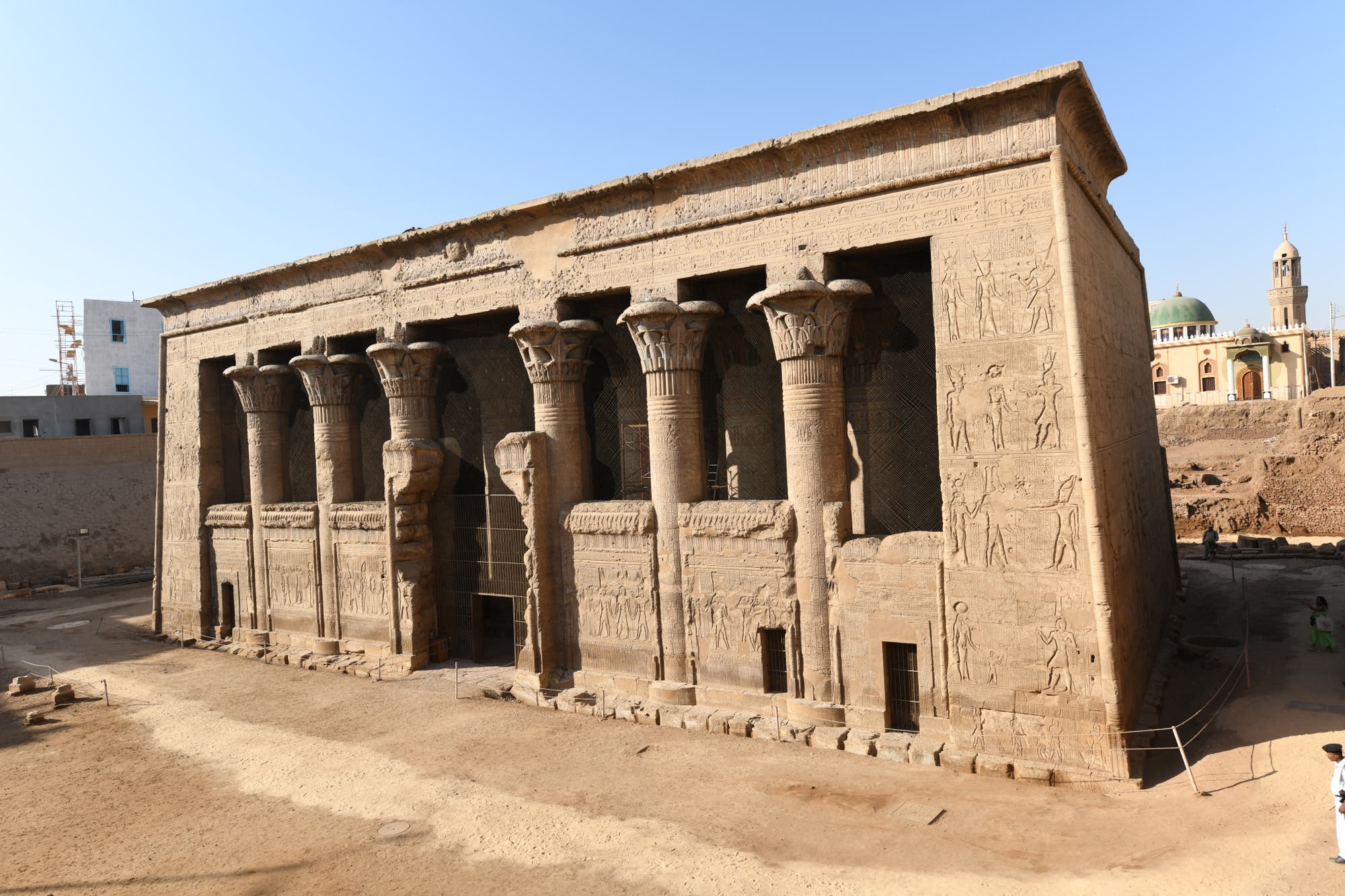 Ein antiker ägyptischer Tempel mit massiven, reich verzierten Säulen und detaillierten Hieroglyphen an den Wänden. Der Tempel steht in einer sandigen Umgebung, umgeben von modernen Gebäuden im Hintergrund. Die Architektur zeigt klassische Merkmale der altägyptischen Baukunst. Keine Personen sind im Bild zu sehen.