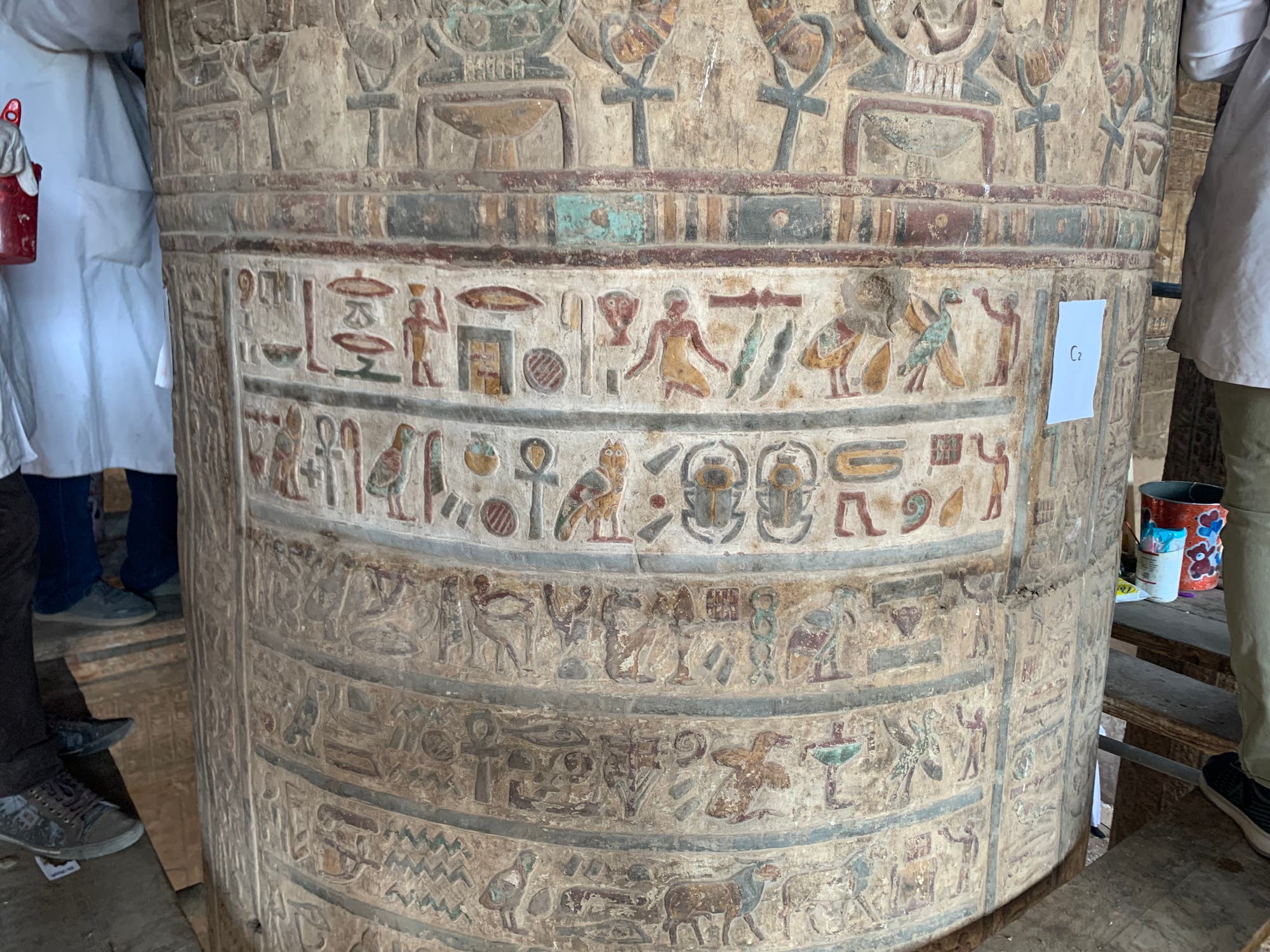 Eine antike Säule mit detaillierten ägyptischen Hieroglyphen und farbigen Symbolen. Zwei Personen in weißen Kitteln stehen links und rechts der Säule, bei der Restaurierung. Ein Blatt Papier mit der Aufschrift "G2" ist an der Säule befestigt. Im Hintergrund sind Werkzeuge und ein Eimer zu sehen.