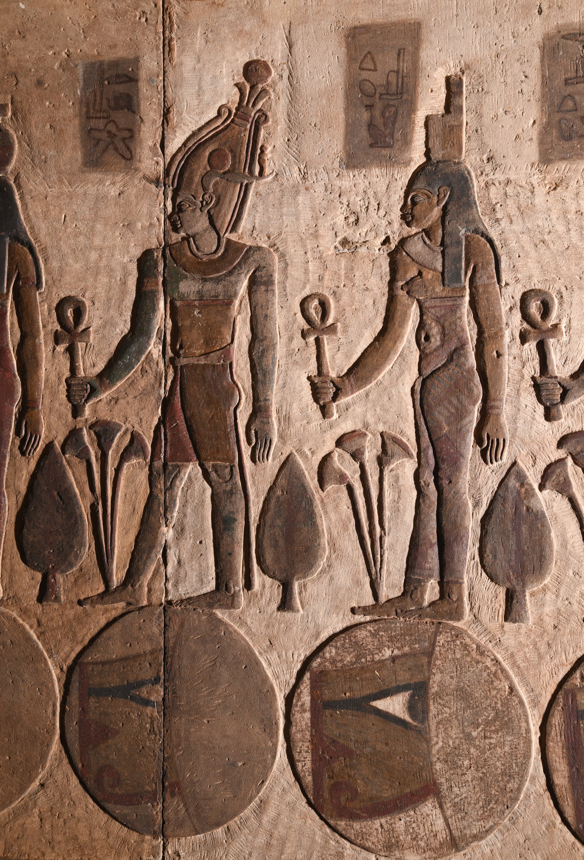 Eine antike ägyptische Wandreliefdarstellung zeigt zwei Figuren, die Ankh-Symbole halten. Beide Figuren tragen traditionelle Kopfbedeckungen und stehen zwischen stilisierten Pflanzenmotiven. Über ihnen sind Hieroglyphen aufgemalt. Unter ihnen sind Scheiben mit Augen darin dargestellt. Die Szene ist in erdigen Tönen gehalten und zeigt typische Merkmale der ägyptischen Kunst.