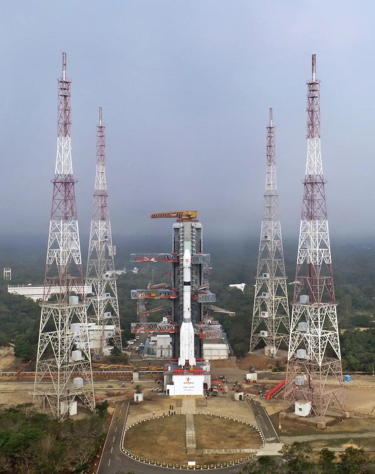 Eine Rakete steht auf einer Startrampe, umgeben von vier hohen, rot-weißen Sendemasten. Die Rakete ist bereit für den Start, mit mehreren Plattformen, die sie umgeben. Im Hintergrund sind Bäume und ein bewölkter Himmel zu sehen. Am unteren Rand der Rakete befindet sich ein Logo mit einem roten Pfeil und dem Text "ISRO". Die Szene vermittelt den Eindruck von technischer Vorbereitung und Raumfahrt.