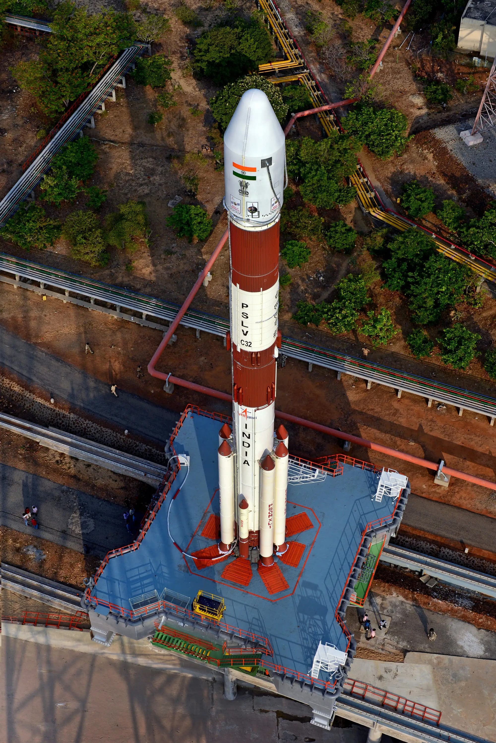 Eine PSLV-Rakete (Polar Satellite Launch Vehicle) steht auf einer Startrampe, bereit für den Start. Die Rakete ist weiß mit roten Akzenten und trägt die Aufschrift "PSLV C32" und "INDIA". Auf der Spitze ist die indische Flagge zu sehen. Die Startrampe ist von Vegetation umgeben, und mehrere Rohre verlaufen parallel zur Rampe. Im Hintergrund sind einige Menschen zu sehen, die sich auf dem Gelände bewegen.