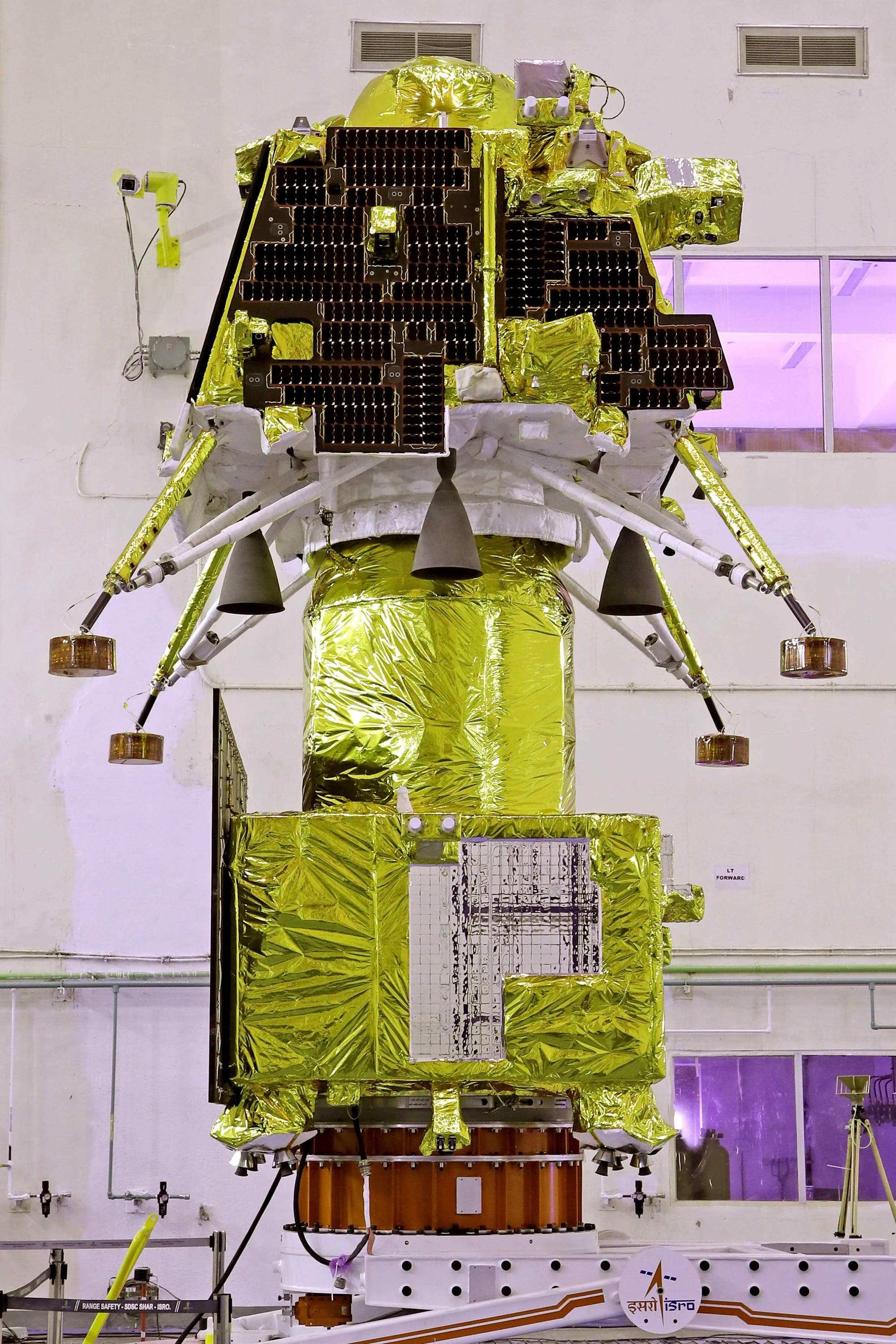 Ein mit goldener Folie umhüllter Satellit in einem Laborraum, der für den Weltraumstart vorbereitet wird. Der Satellit ist mit mehreren Solarpaneelen und Düsen ausgestattet. Im Hintergrund sind violett beleuchtete Fenster und technische Ausrüstung zu sehen. Unten ist das Logo der indischen Weltraumorganisation ISRO erkennbar.