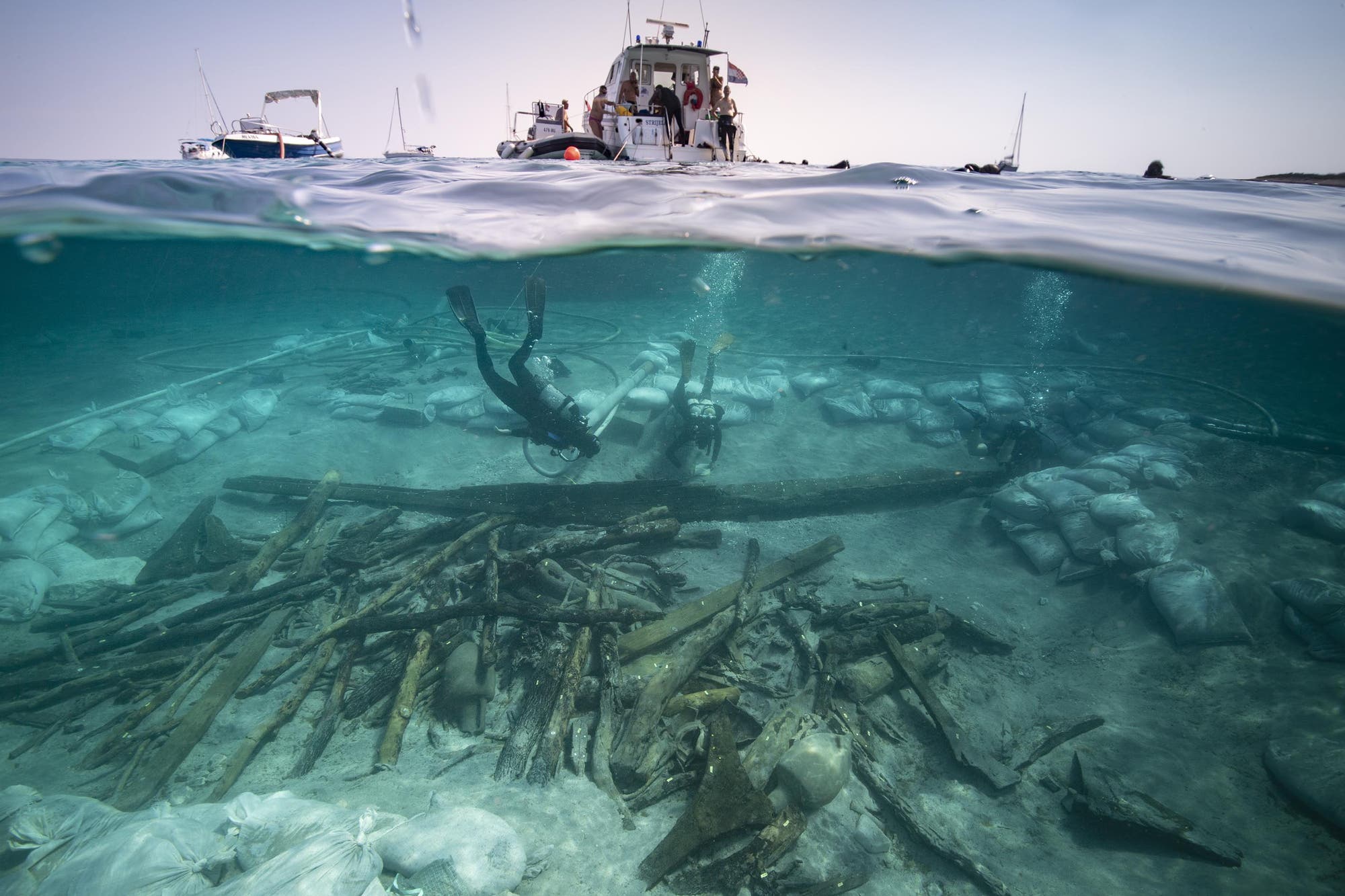 Taucher erkunden eine Unterwasser-Ausgrabungsst&auml;tte mit Holztr&uuml;mmern, Amphoren und Sands&auml;cken auf dem Meeresboden. &Uuml;ber der Wasseroberfl&auml;che ist ein Boot mit mehreren Personen zu sehen. Die Szene zeigt eine arch&auml;ologische Untersuchung in einem klaren, blauen Meer.
